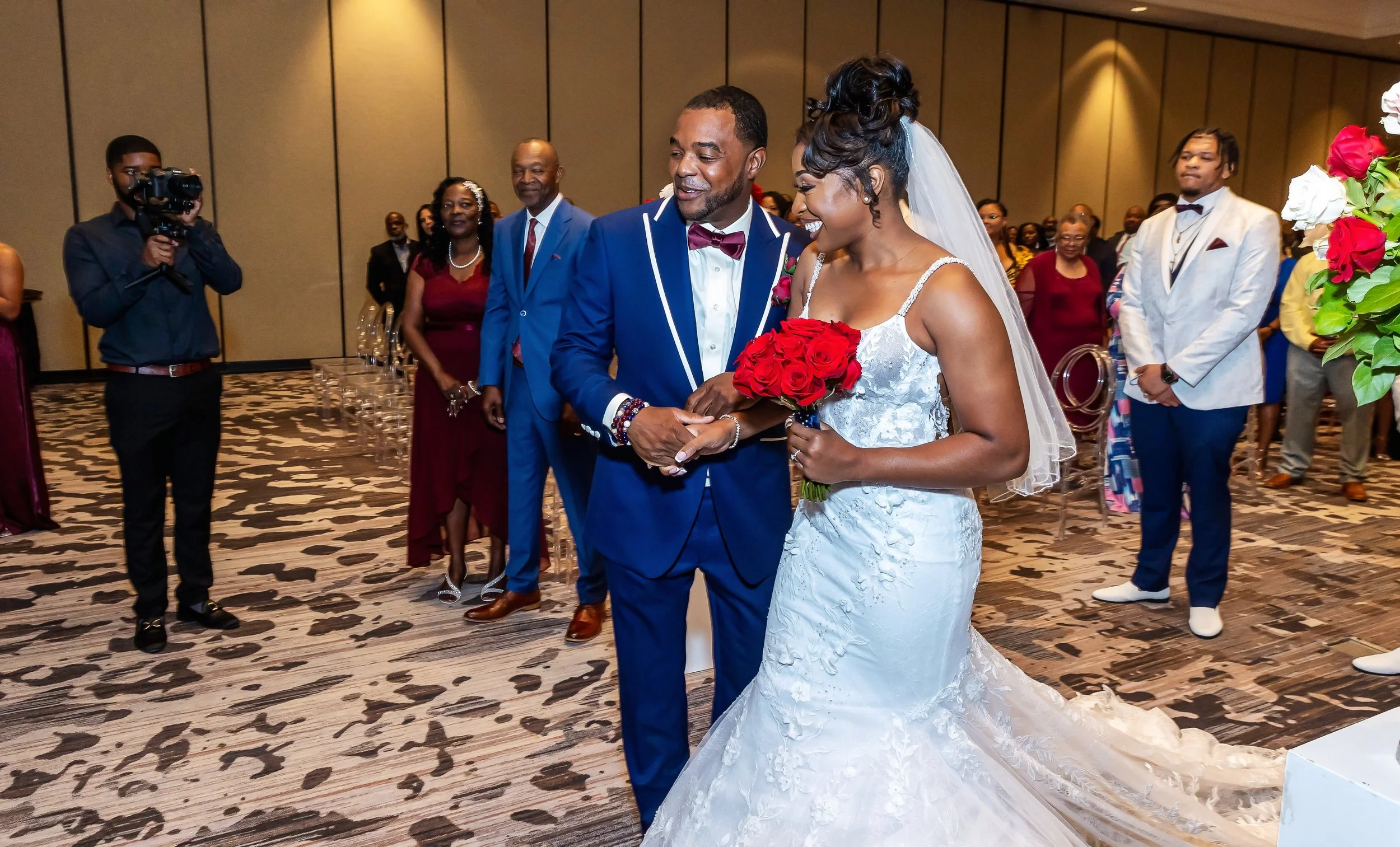 A bride and groom at their wedding ceremony, holding hands and smiling, surrounded by guests, with beautiful floral arrangements in the background.