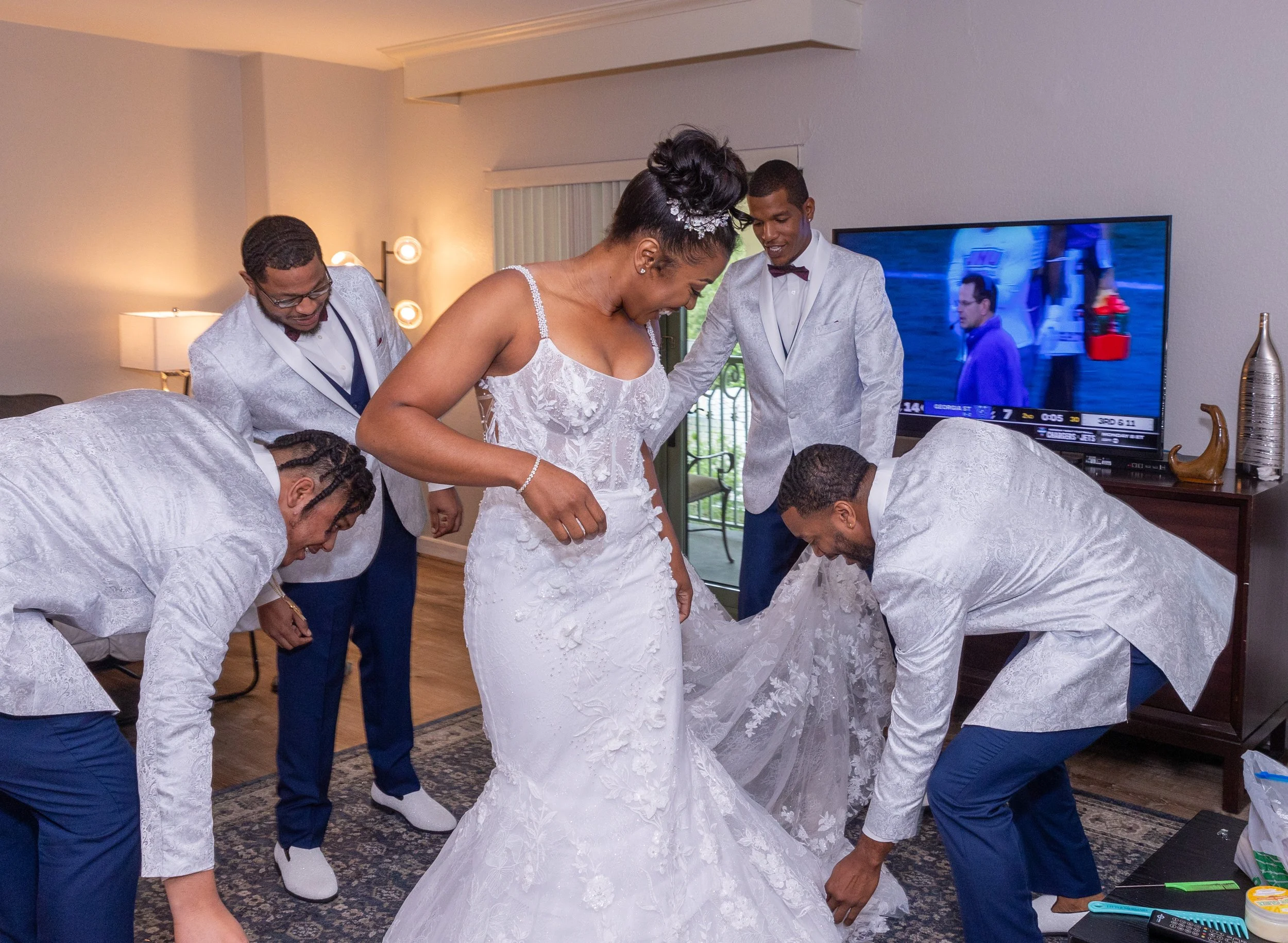 A bride in a white wedding dress smiling as she stands among five men in suits, who are helping her prepare for the wedding inside a room with a TV and decorative objects.