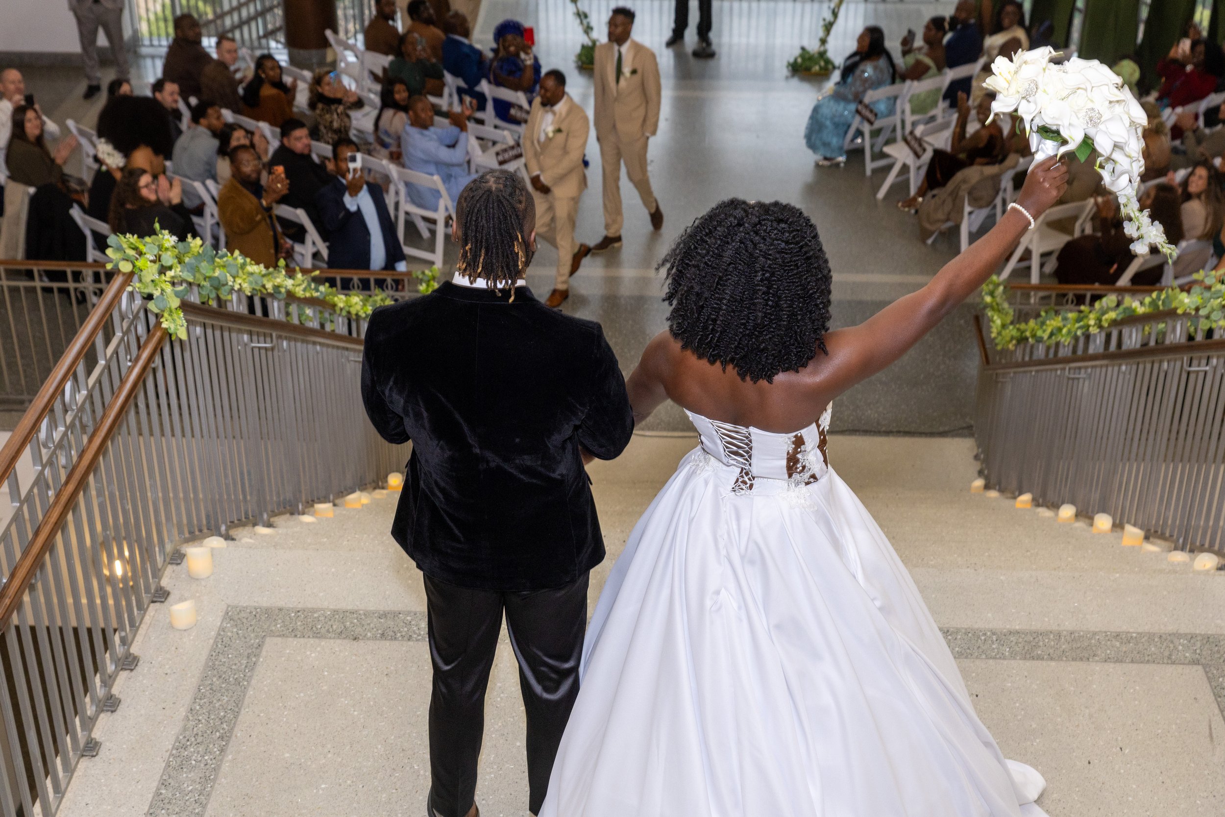 A bride and groom walking down the stairs at their wedding ceremony, with wedding guests seated and standing on either side, some taking photos. The bride holds a bouquet of white flowers, and the groom is dressed in a black tuxedo. The setting is in