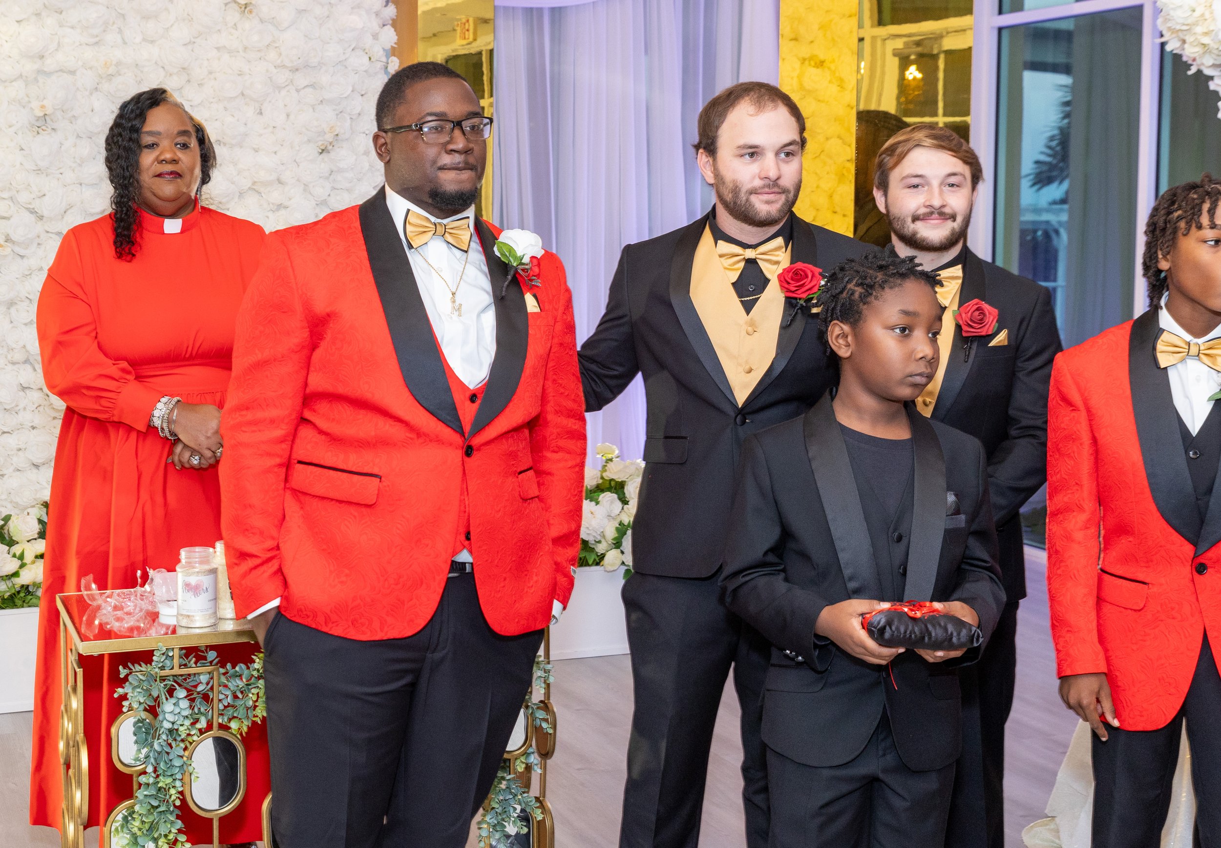 Group of diverse people dressed formally at a celebration or ceremony, with dressed attire including tuxedos and dresses, in a decorated indoor venue.
