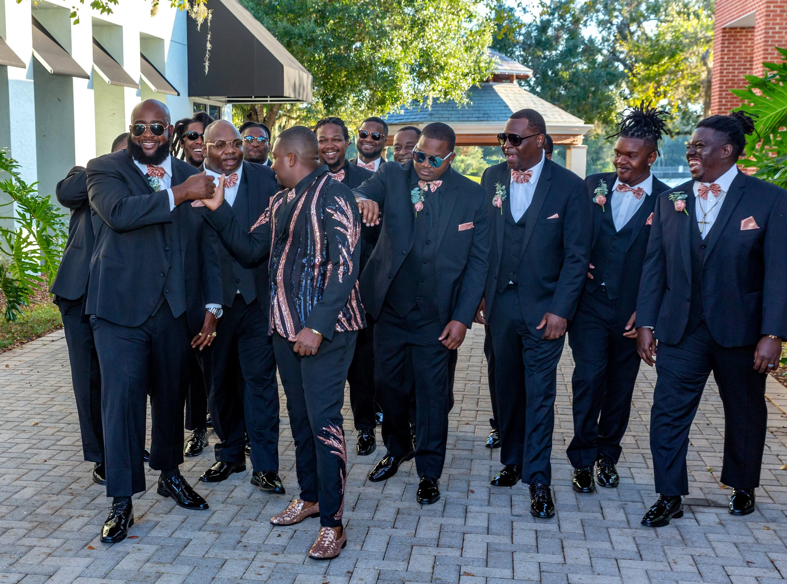 A group of men dressed in black tuxedos with pink bow ties and boutonnières, walking outdoors on a paved path during a celebration, with one woman in the front wearing a sequin outfit.