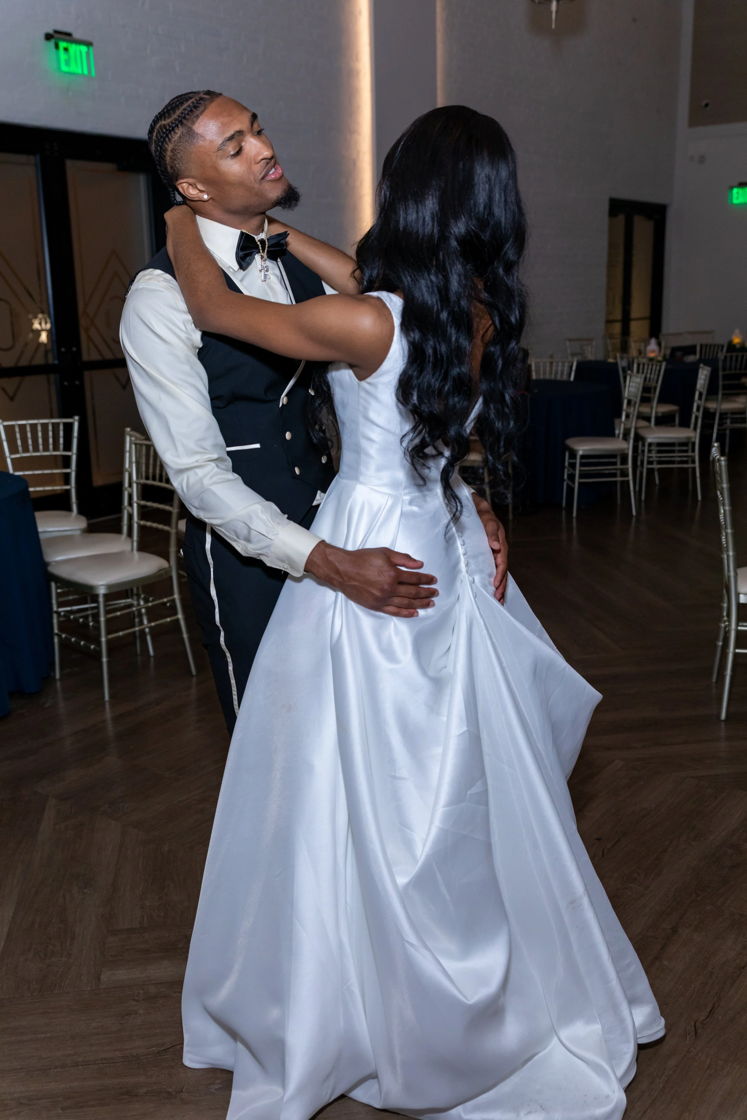 A man and woman dancing together in a celebration, with the man wearing formal attire including a bow tie and vest, and the woman wearing a white gown, in a venue with chairs and tables in the background.
