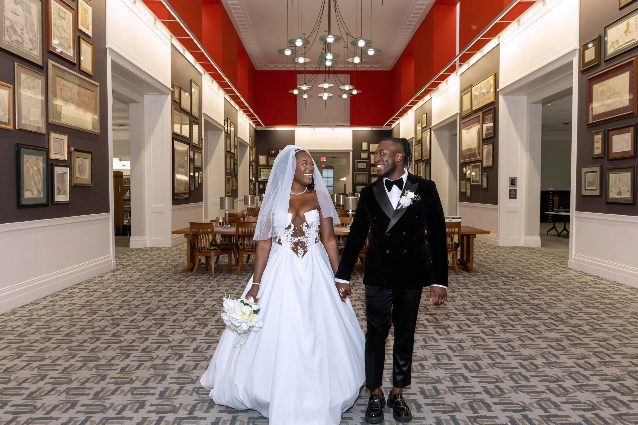 A bride and groom smile at each other while holding hands, walking through a decorated indoor hall with framed pictures on the walls and patterned carpet.