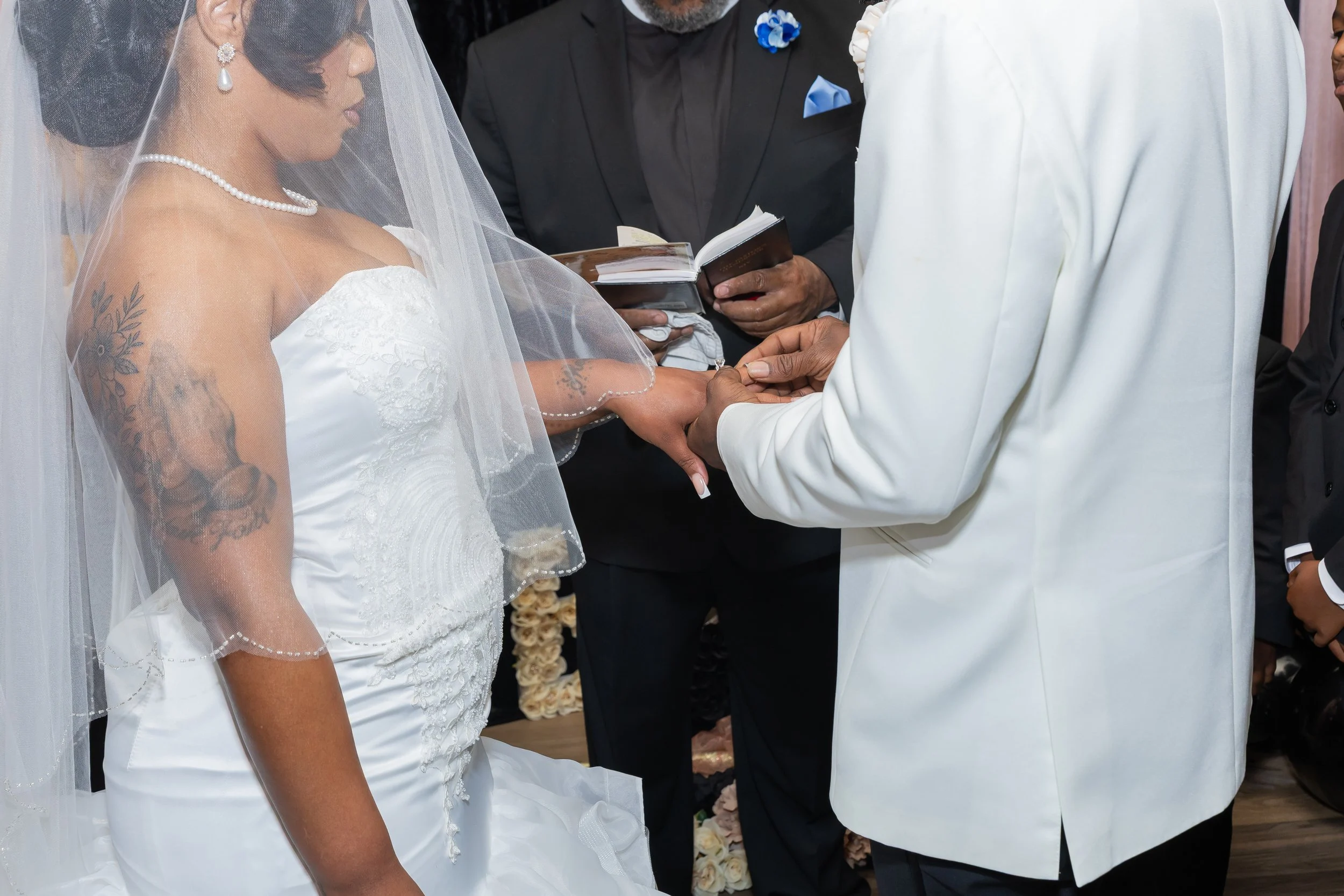 A bride and groom exchanging wedding vows during their ceremony, with an officiant in the background holding a book. The bride wears a white lace wedding dress and veil, and has tattoos on her arm. The groom wears a white suit.
