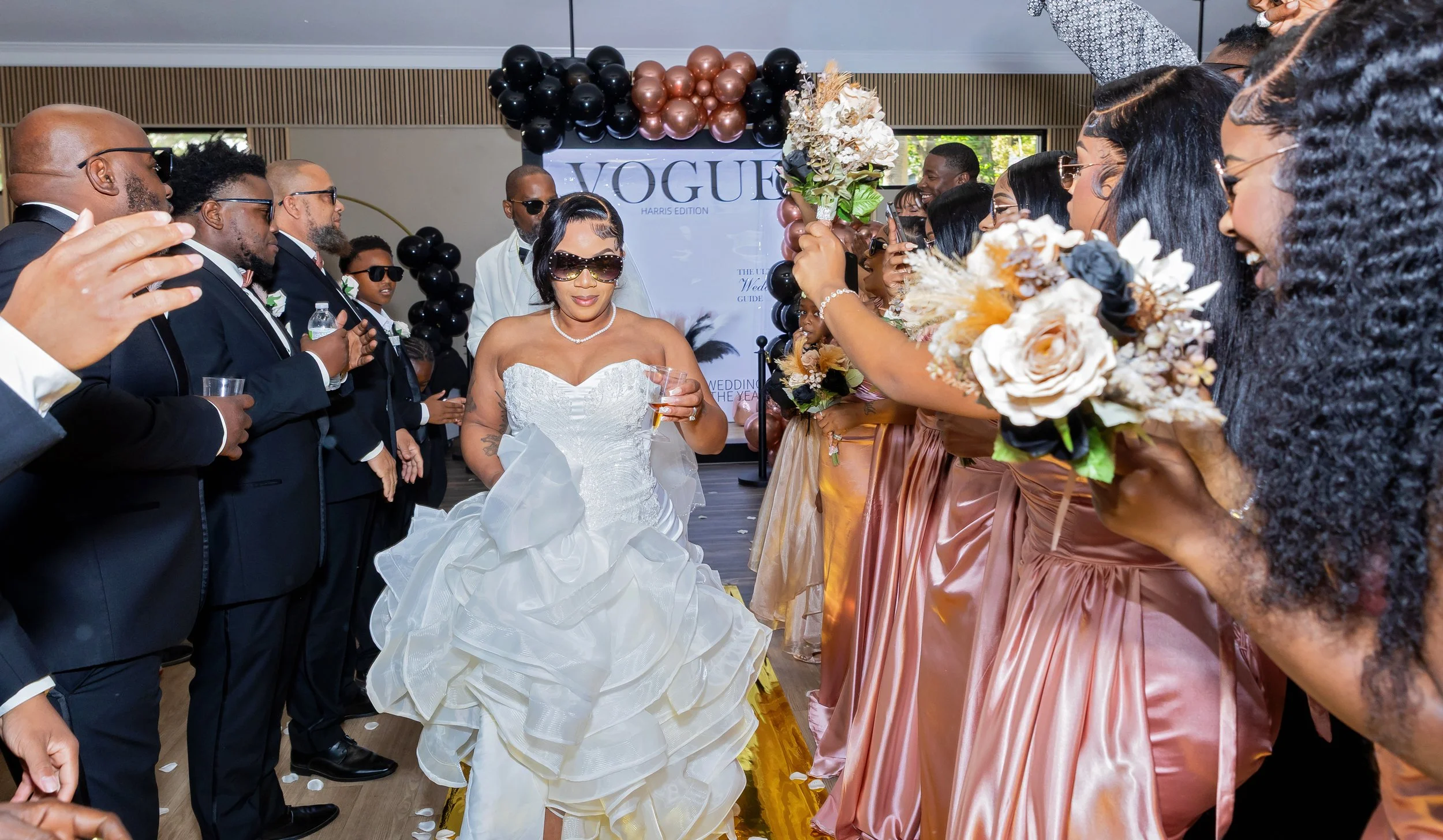 A woman in a white wedding dress and sunglasses walks through a line of women in pink dresses holding bouquets, with a display reading 'VOGUE' and black and brown balloons in the background.
