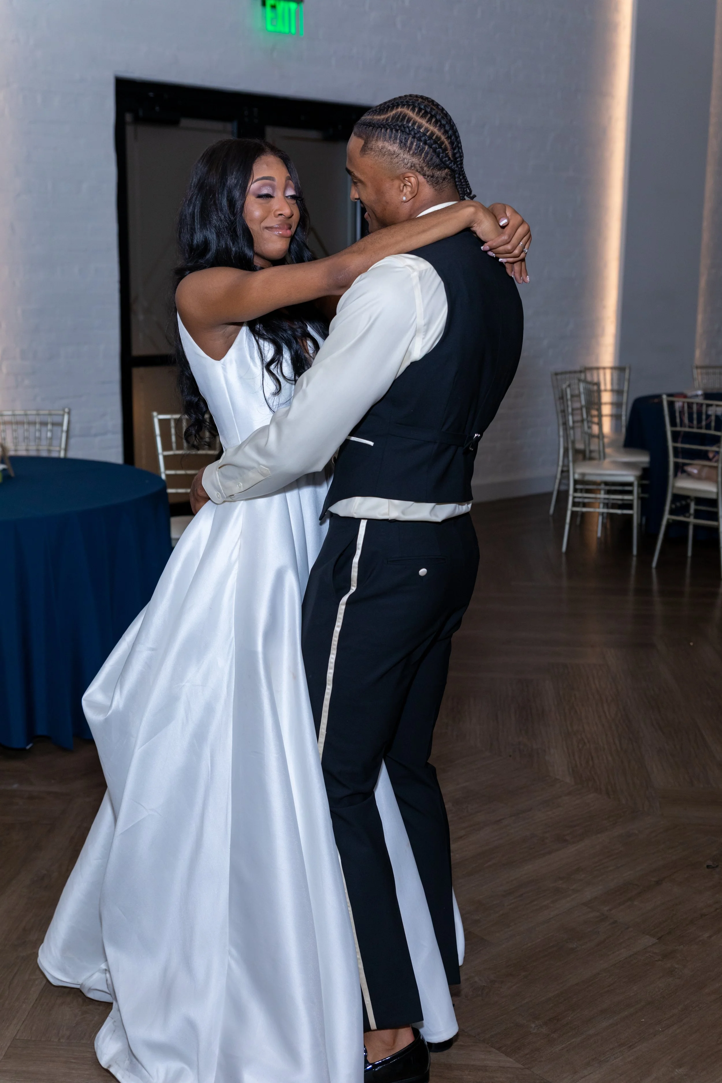 A couple dancing closely at an event, with the woman dressed in a white gown and the man in a black vest and pants.