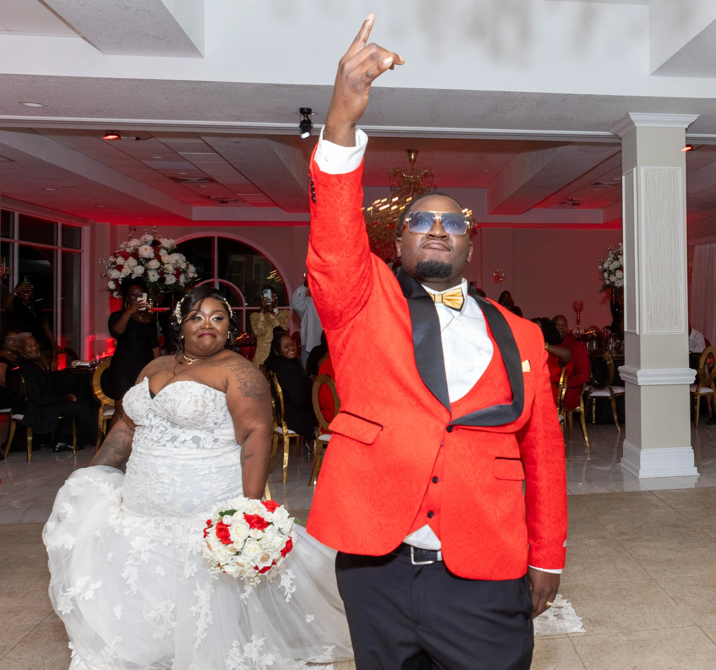 A man in a red tuxedo jacket and sunglasses, with a woman in a wedding dress holding a bouquet of red and white flowers at a wedding reception.