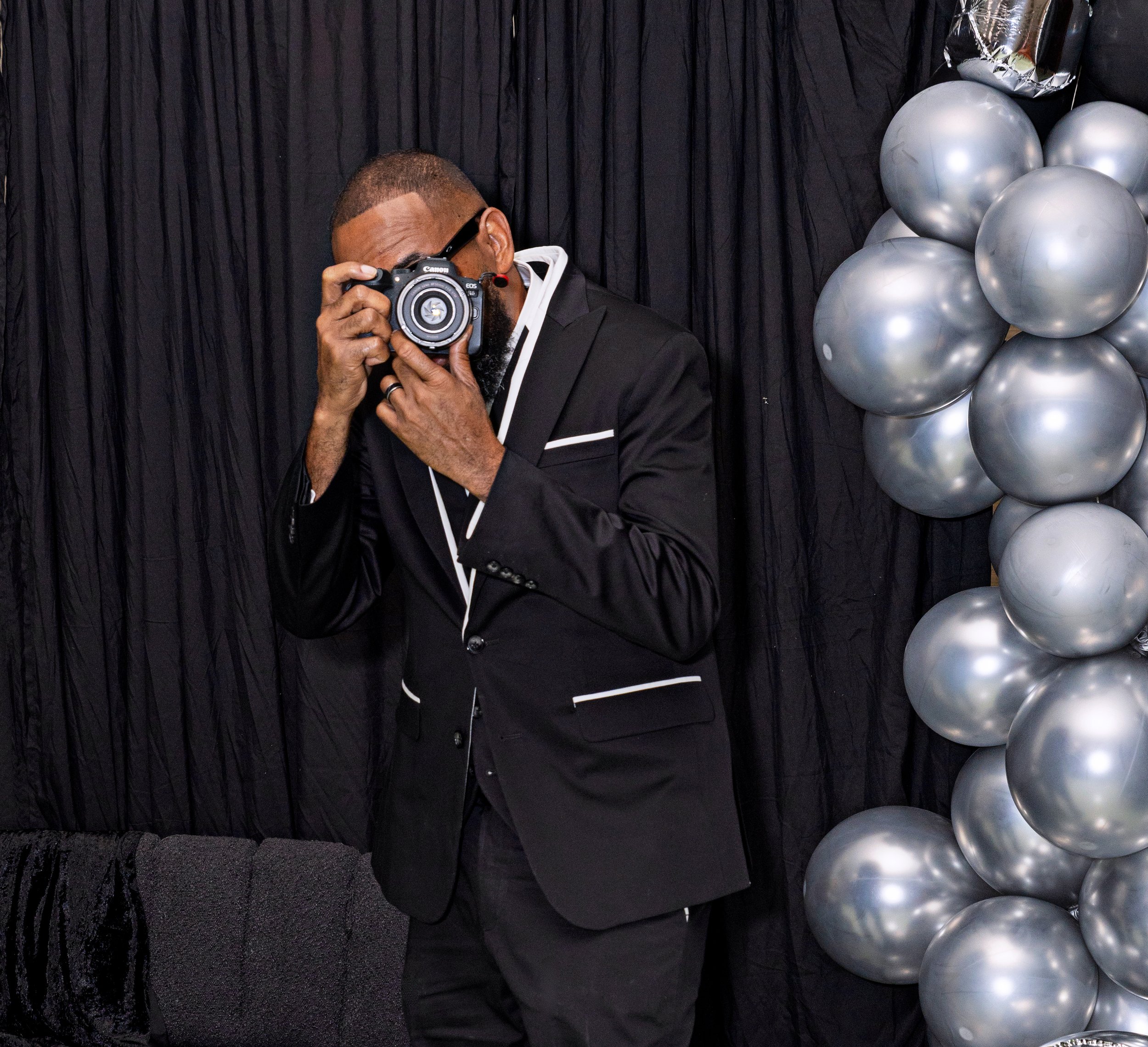 A man in a suit taking a photo with a camera at an event. Black backdrop with black curtains and silver balloons in the background.