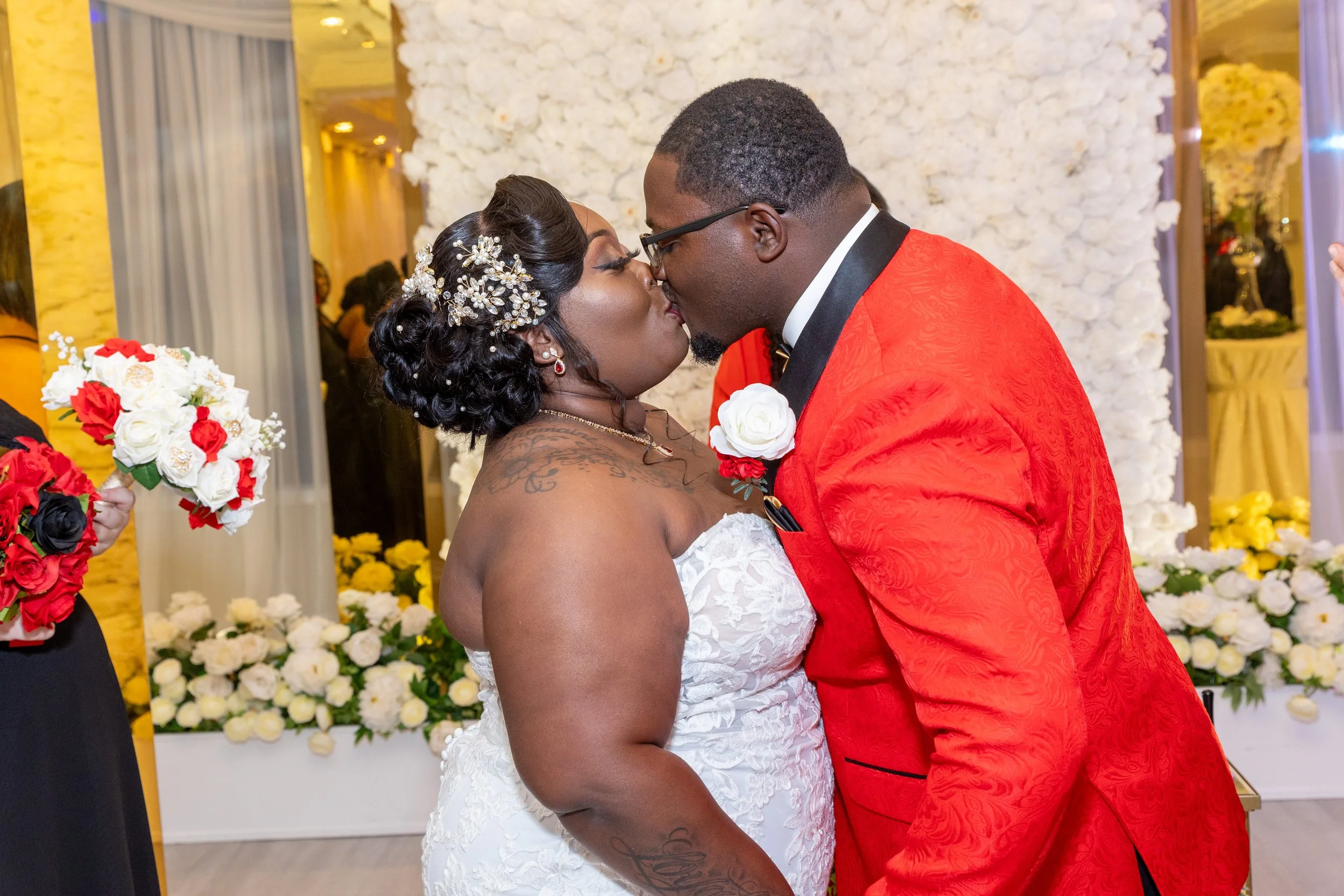 A bride and groom sharing a kiss at their wedding reception, surrounded by floral decorations in white, red, and yellow, with guests in the background.
