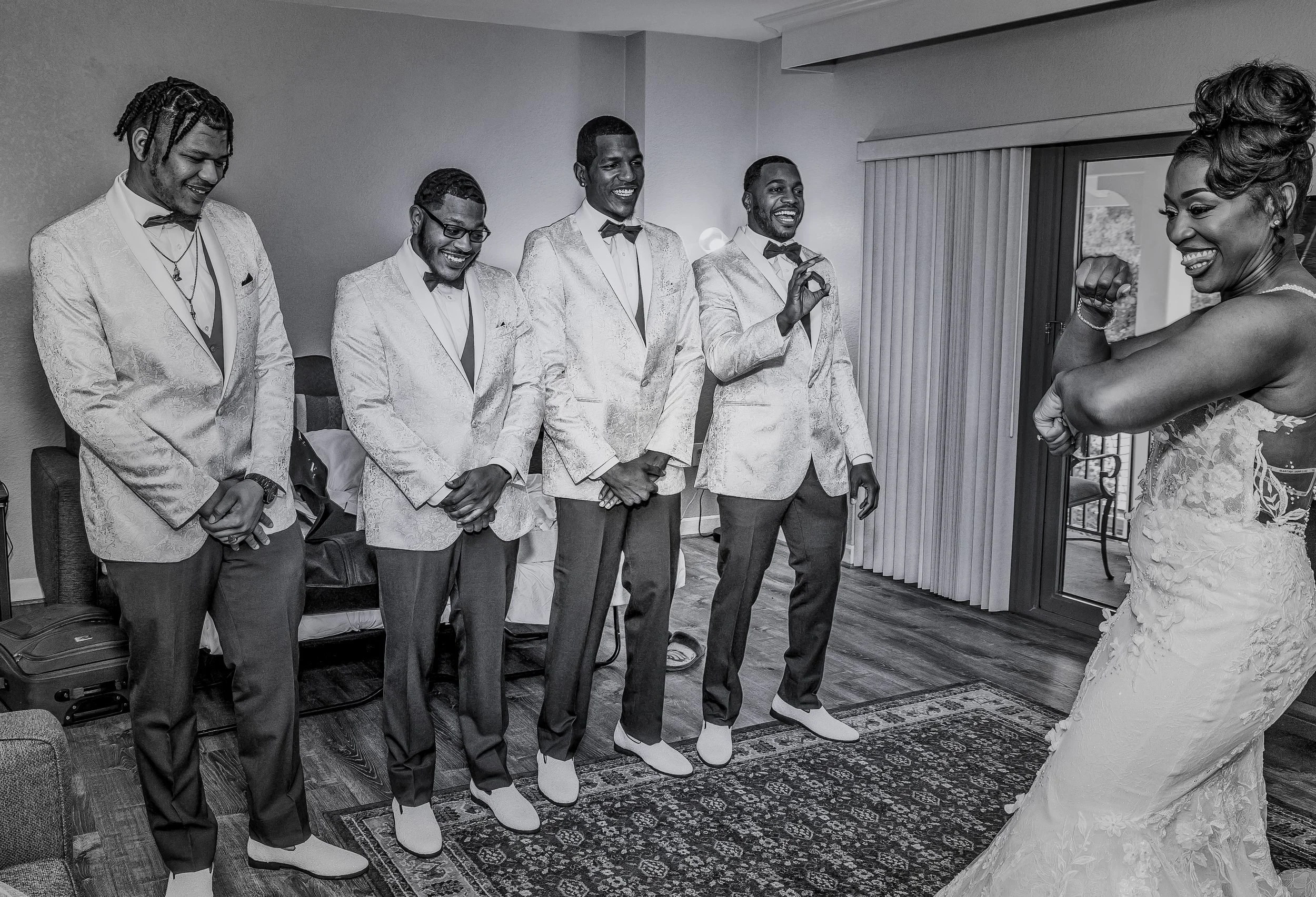 Group of five men dressed in tuxedos with bow ties, smiling and standing in a line, facing a woman in an elegant dress during a wedding celebration.