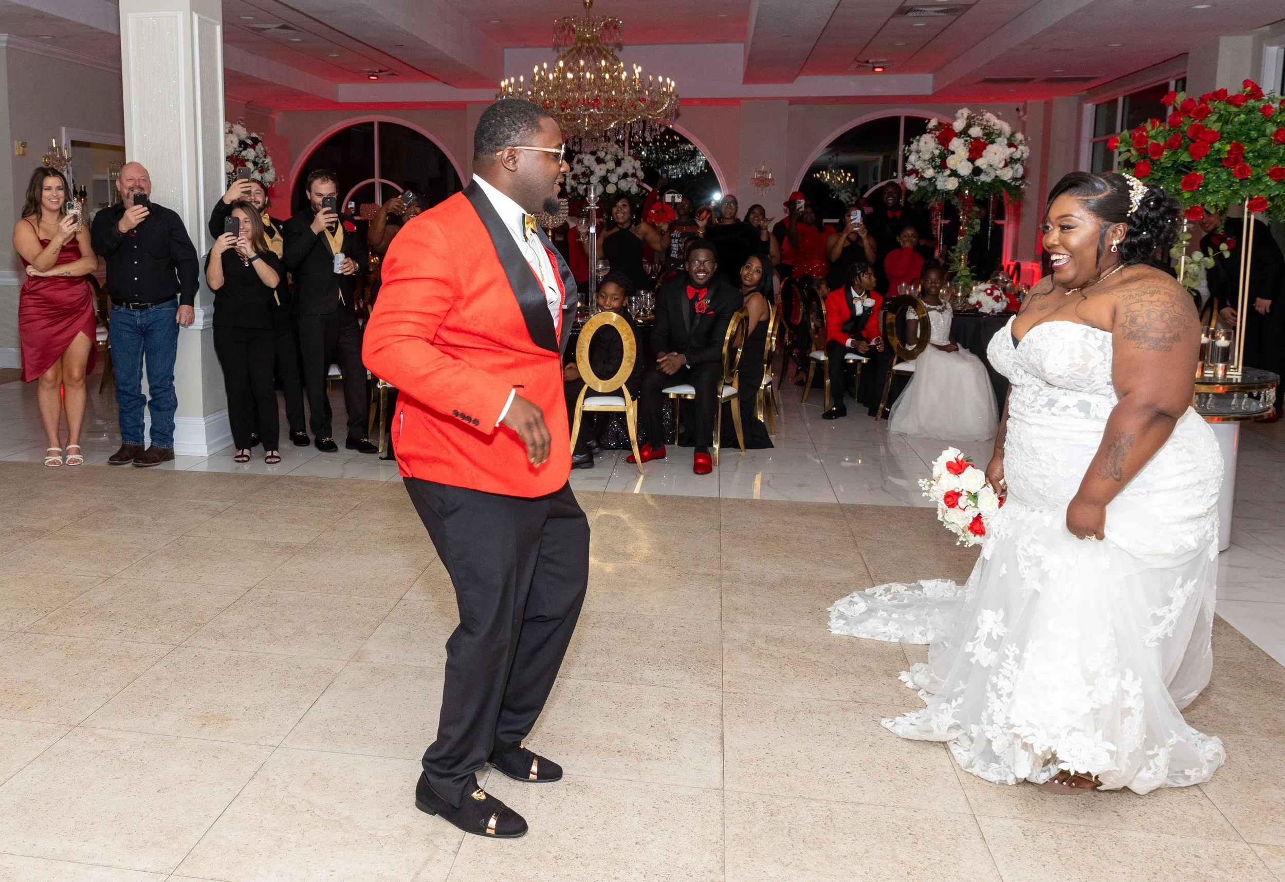 Bride and groom are dancing at their wedding reception, surrounded by guests in formal attire, with large floral arrangements and elegant decorations in the background.