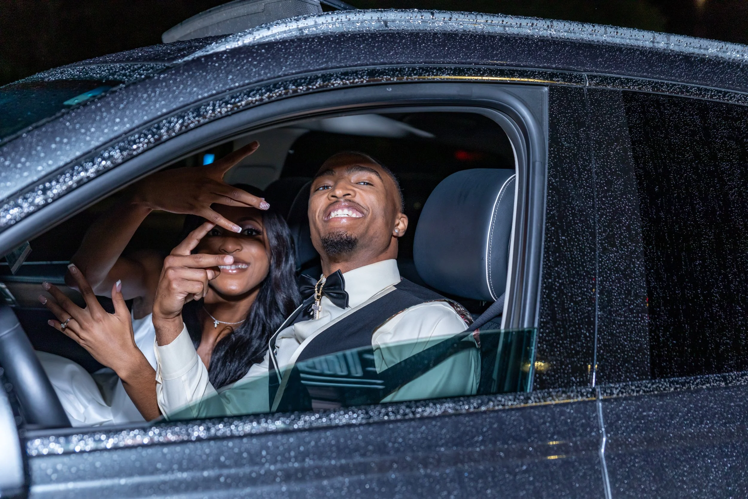 A man and woman dressed in formal attire sitting inside a luxury car, smiling and posing for the camera. The car's exterior is decorated with sparkling black glitter and raindrops.