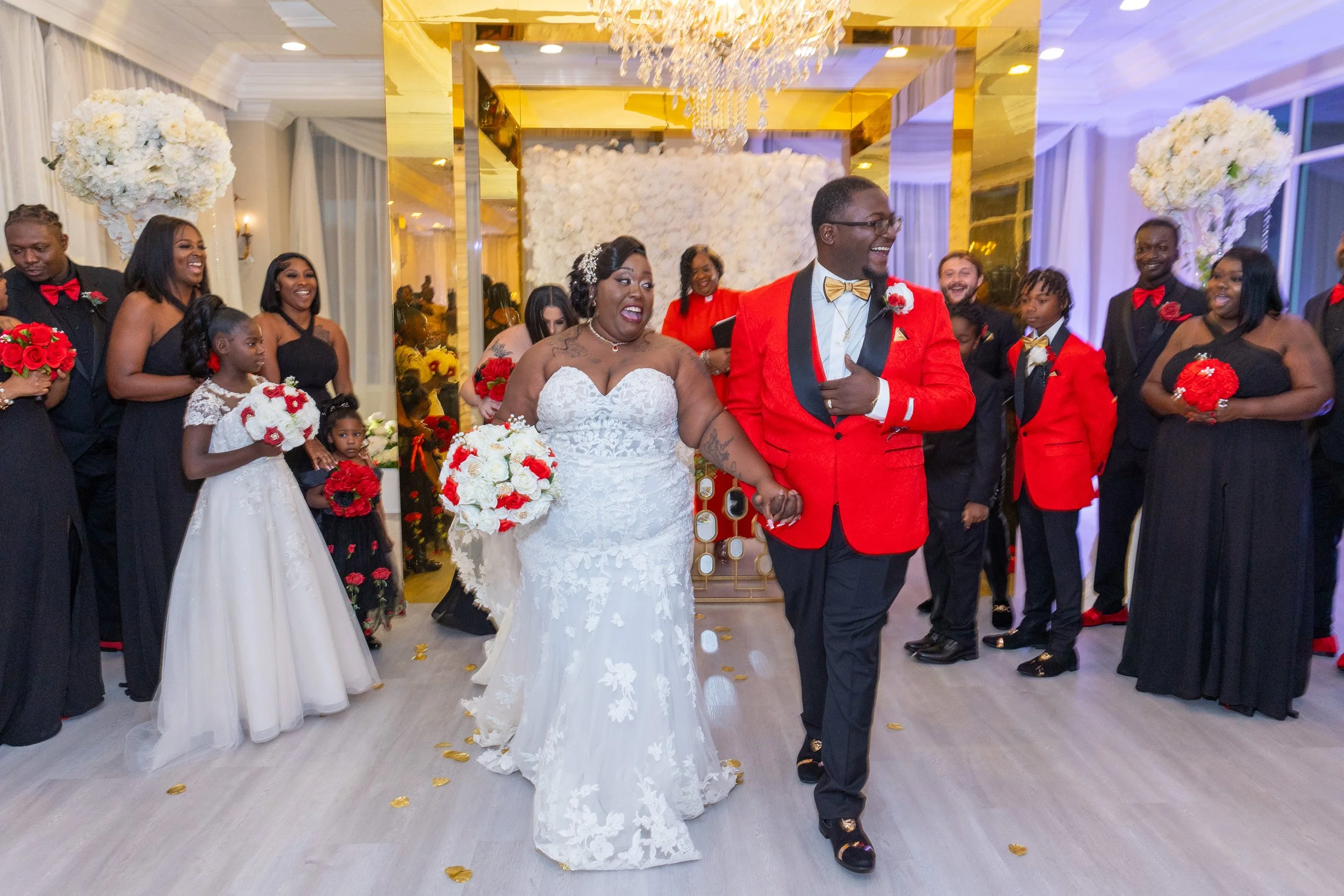 A wedding celebration with a bride and groom holding hands, surrounded by friends and family. The bride wears a white lace wedding dress and carries a bouquet of white and red flowers, while the groom is dressed in a red tuxedo jacket with black acce