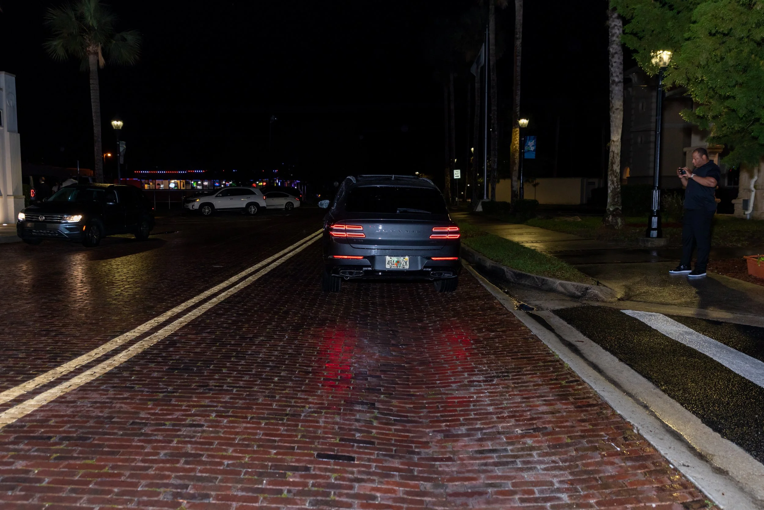Nighttime view of a street with parked cars, streetlights, and a man standing on the sidewalk using his phone.