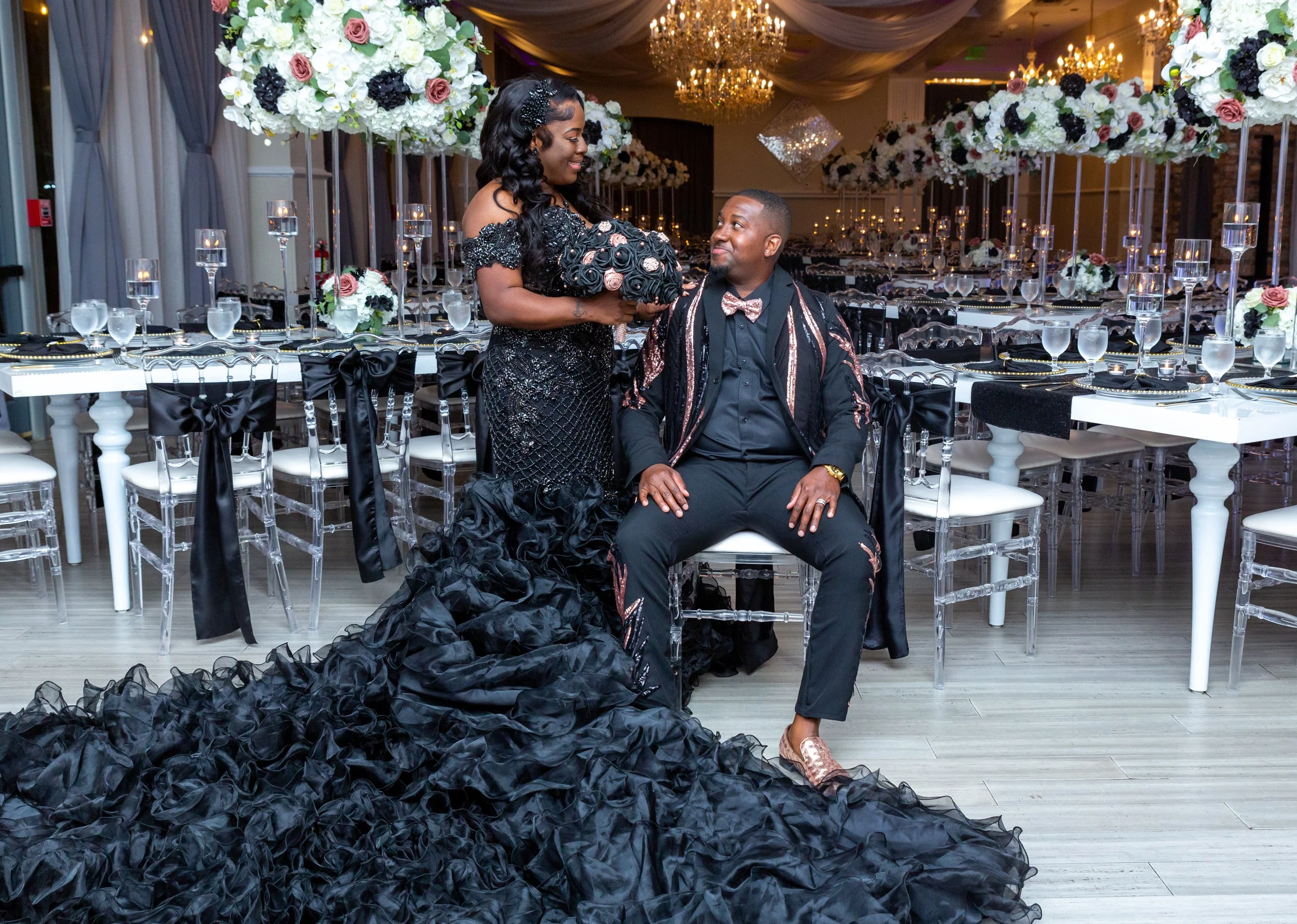 A woman in a black sequined gown holding a bouquet of black and pink roses, smiling at a man seated in a tuxedo with a pink and black bow tie, in an elegantly decorated banquet hall with floral centerpieces and chandeliers.
