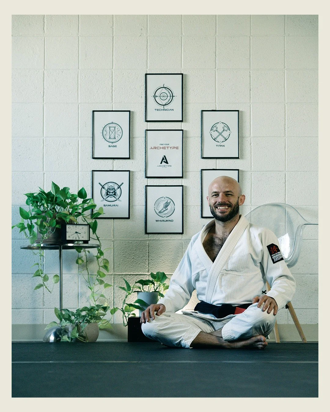 A man in a white martial arts uniform sitting cross-legged on a black mat, smiling, in a room with framed artwork and green plants on a shelf.
