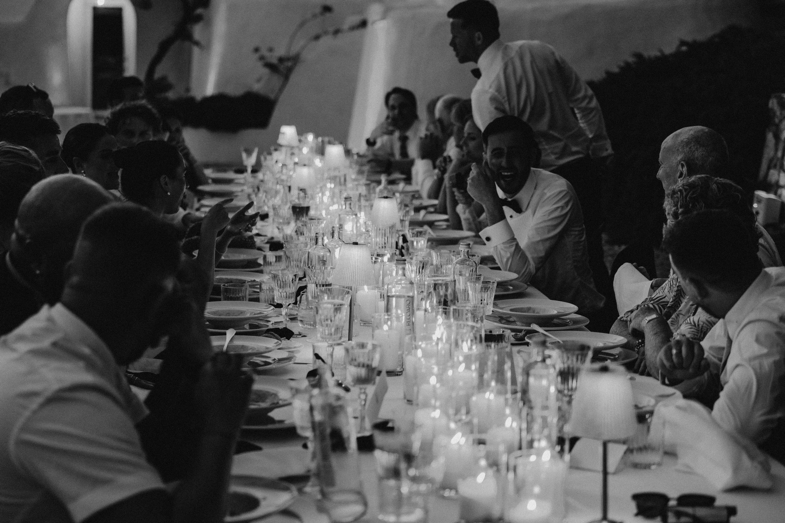 A black and white photograph of a formal dinner party with many people seated around a long table, enjoying conversation. A waiter is serving the guests, and the setting appears elegant with tableware and glasses.