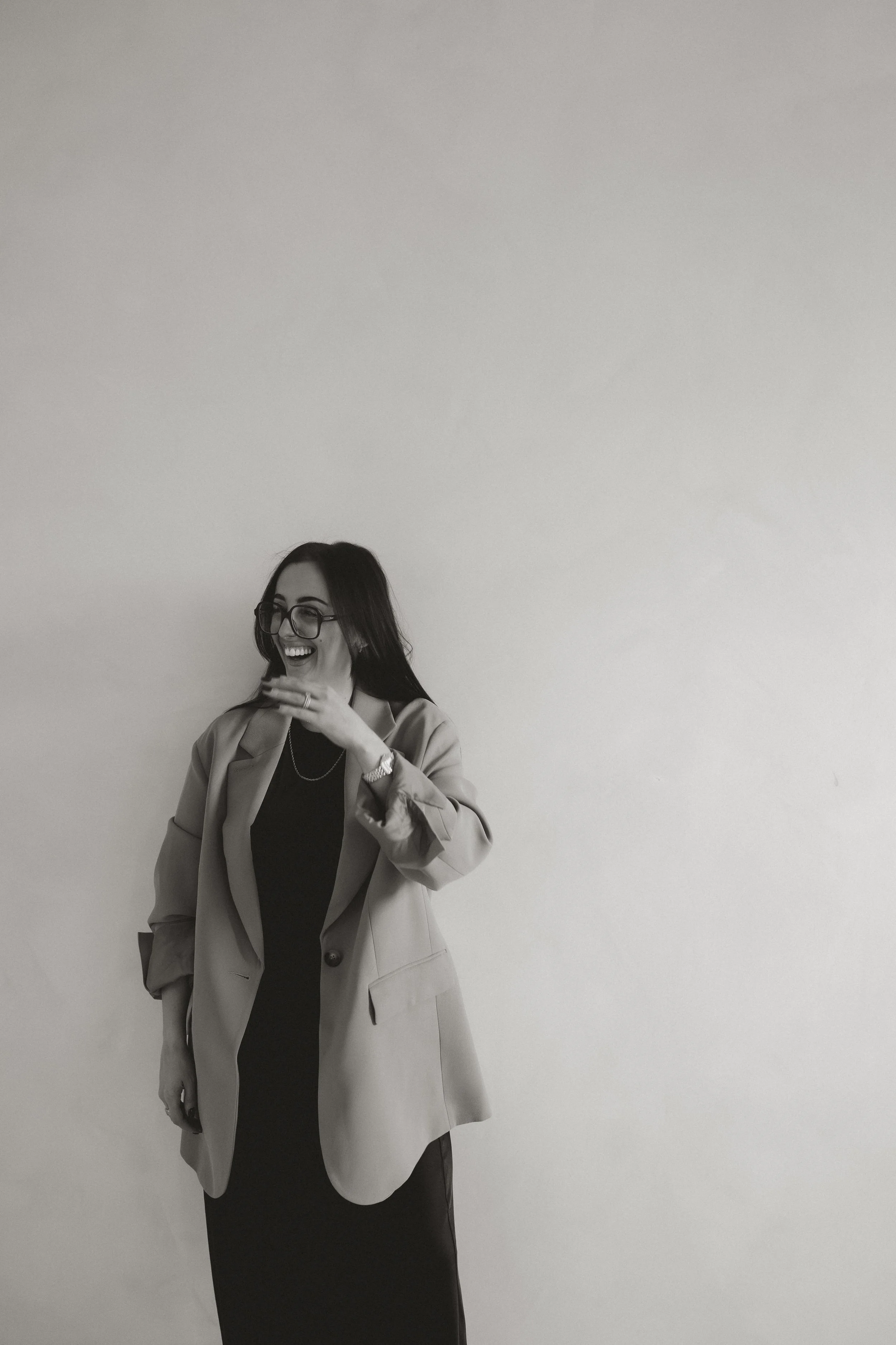 A woman with glasses and dark hair laughing and smiling, dressed in a blazer and long dress, standing against a plain wall.
