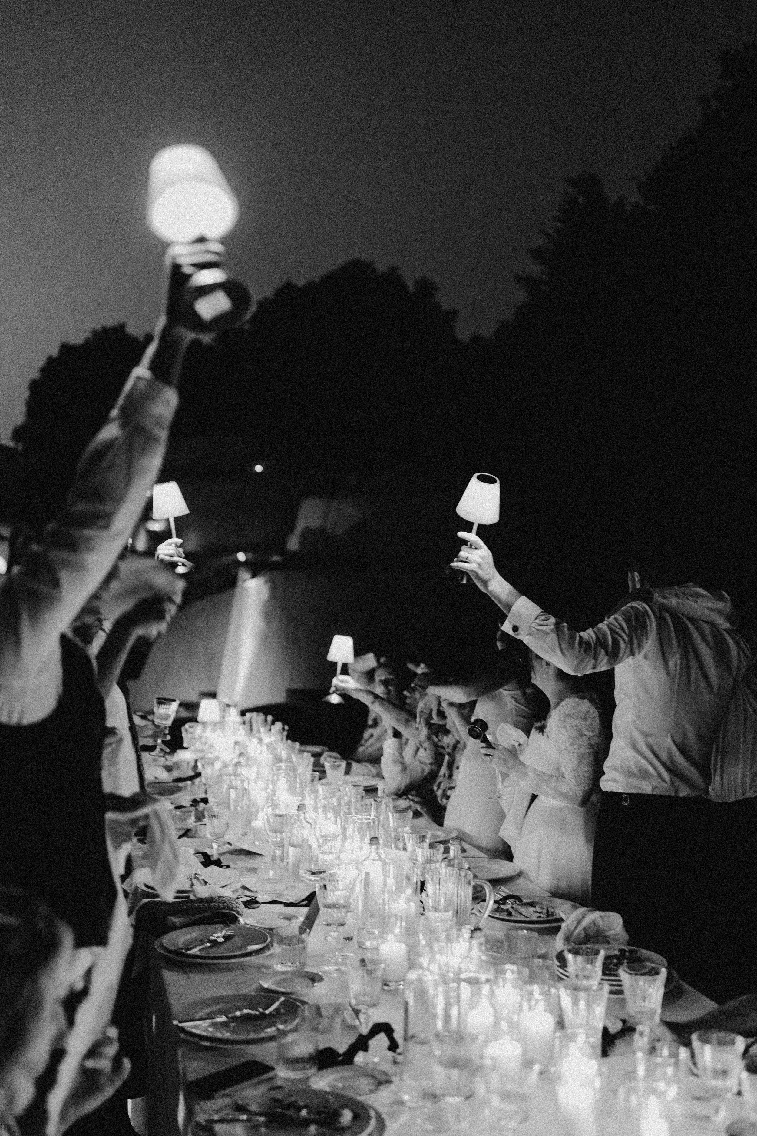 Black and white photo of people at a long table holding small lamps in the air during an outdoor nighttime gathering.