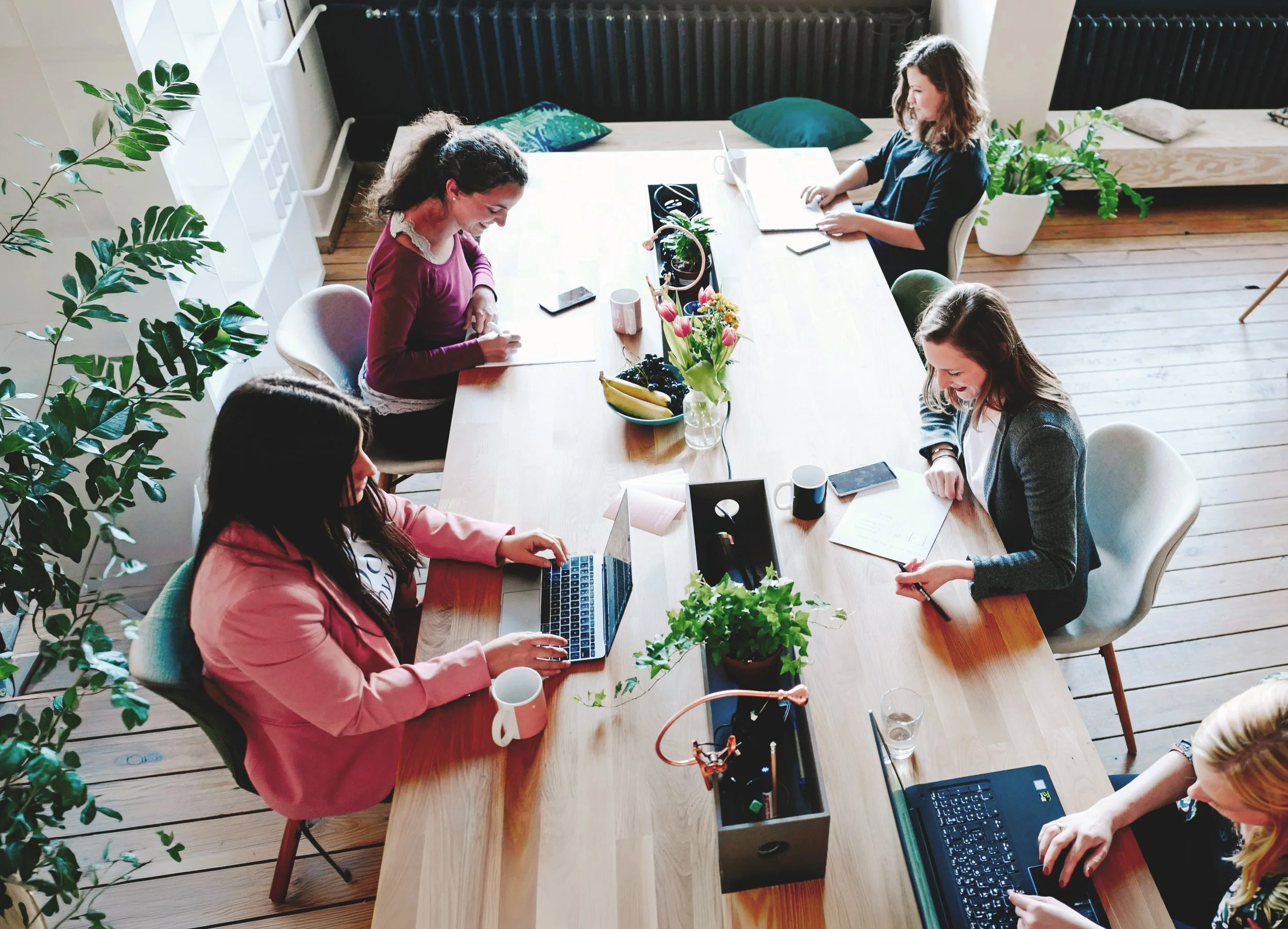 A group of five women gathered around a long wooden table in a bright room with wooden floors. They are engaged in working on laptops, writing, and talking. The table has a laptop, a tablet, a smartphone, notebooks, pens, and cups. There are potted plants and a bowl of fruit on the table, and a bed with pillows and a large plant in the background.