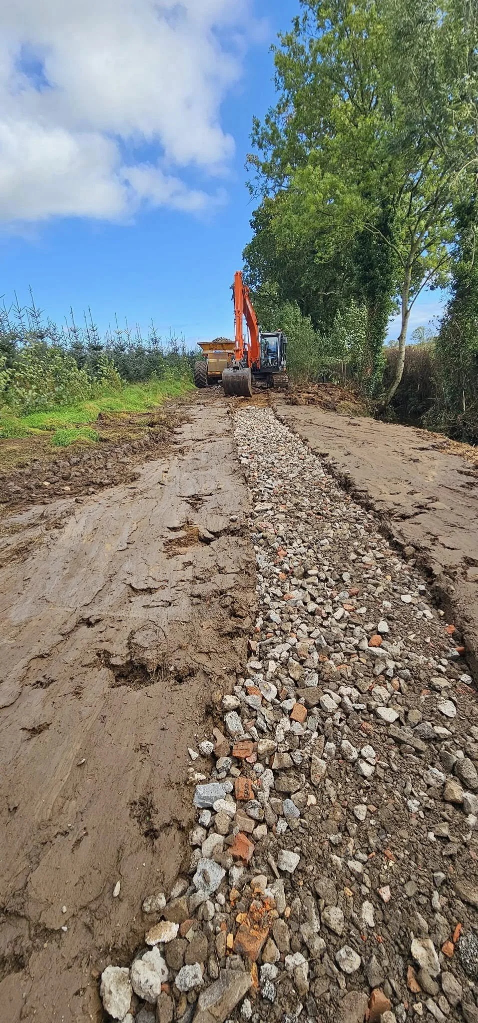 A construction site showing a backhoe loader spreading gravel over the earth for road construction, with trees and blue sky in the background.