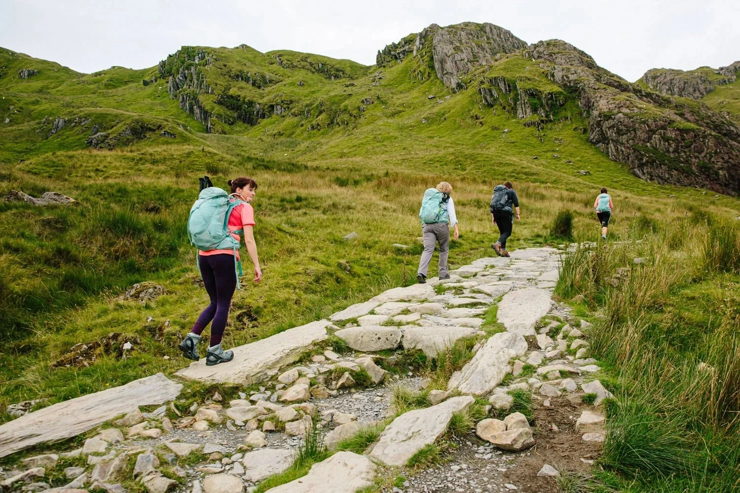 Four hikers with backpacks walking along a rocky trail in a green mountainous landscape.