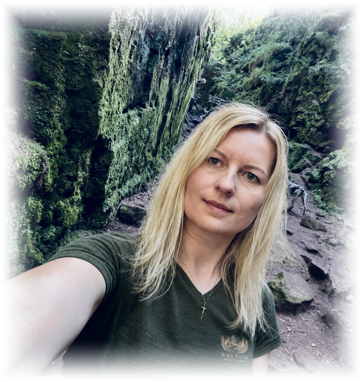 A woman with blonde hair taking a selfie outdoors in a mossy, rocky area of a forest.