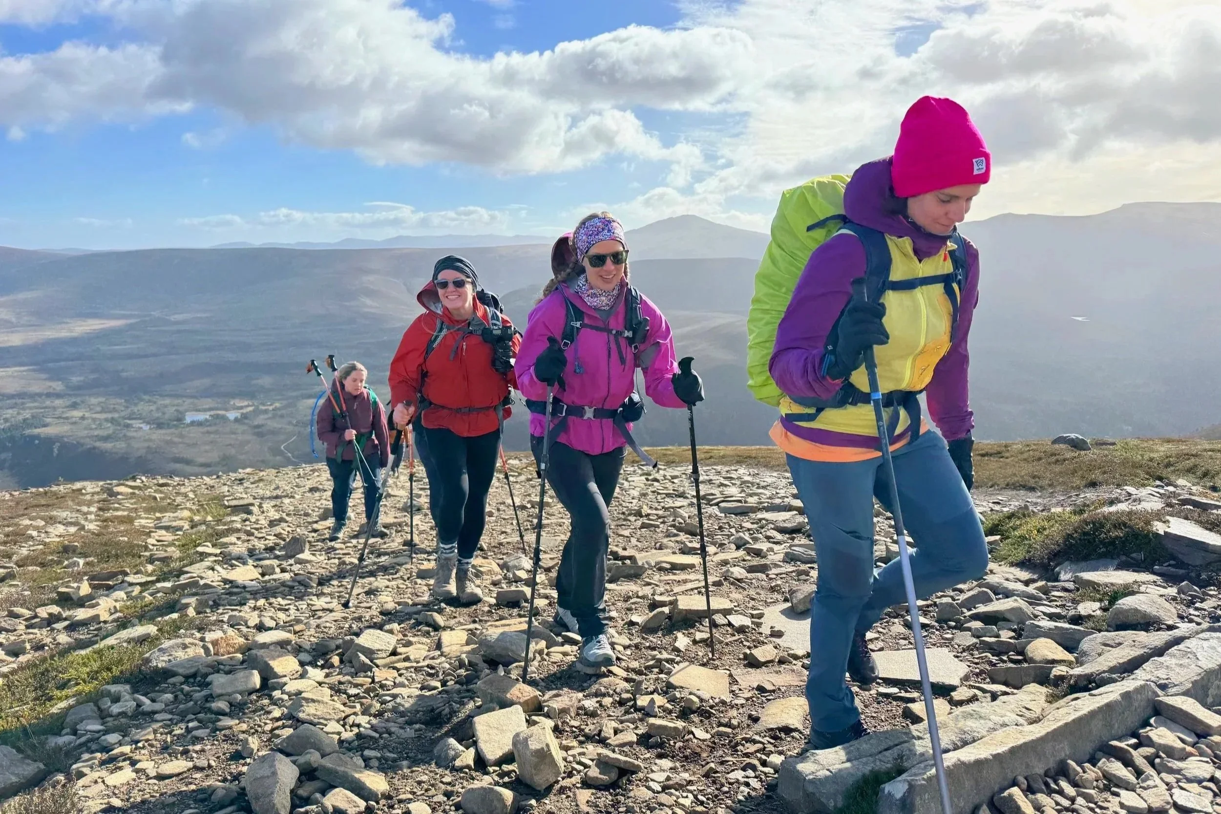 A group of five women hiking in a mountainous area with rocky terrain and cloudy sky, all wearing colorful outdoor gear and carrying backpacks.