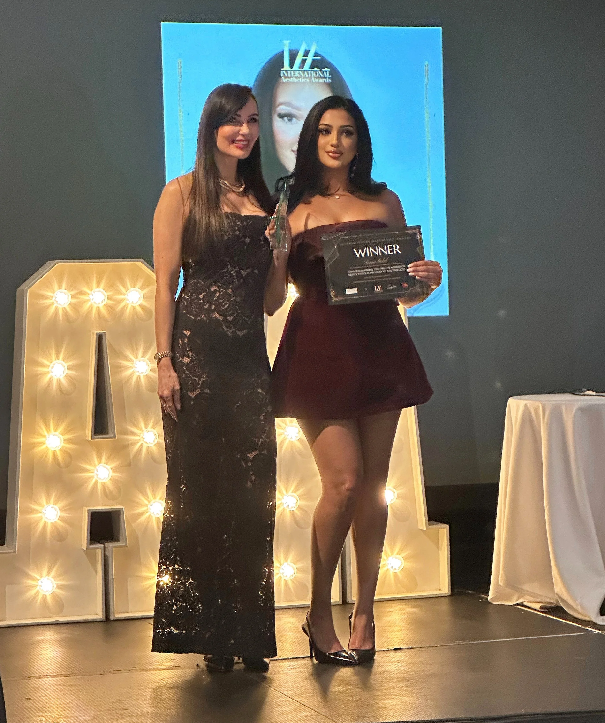 Two women standing on stage at an awards ceremony, one holding a winner plaque. The woman on the left wears a black lace dress; the woman on the right wears a strapless burgundy dress and high heels. Behind them is a large illuminated marquee letter and a blue screen with a face and the words "International Teen Awards."