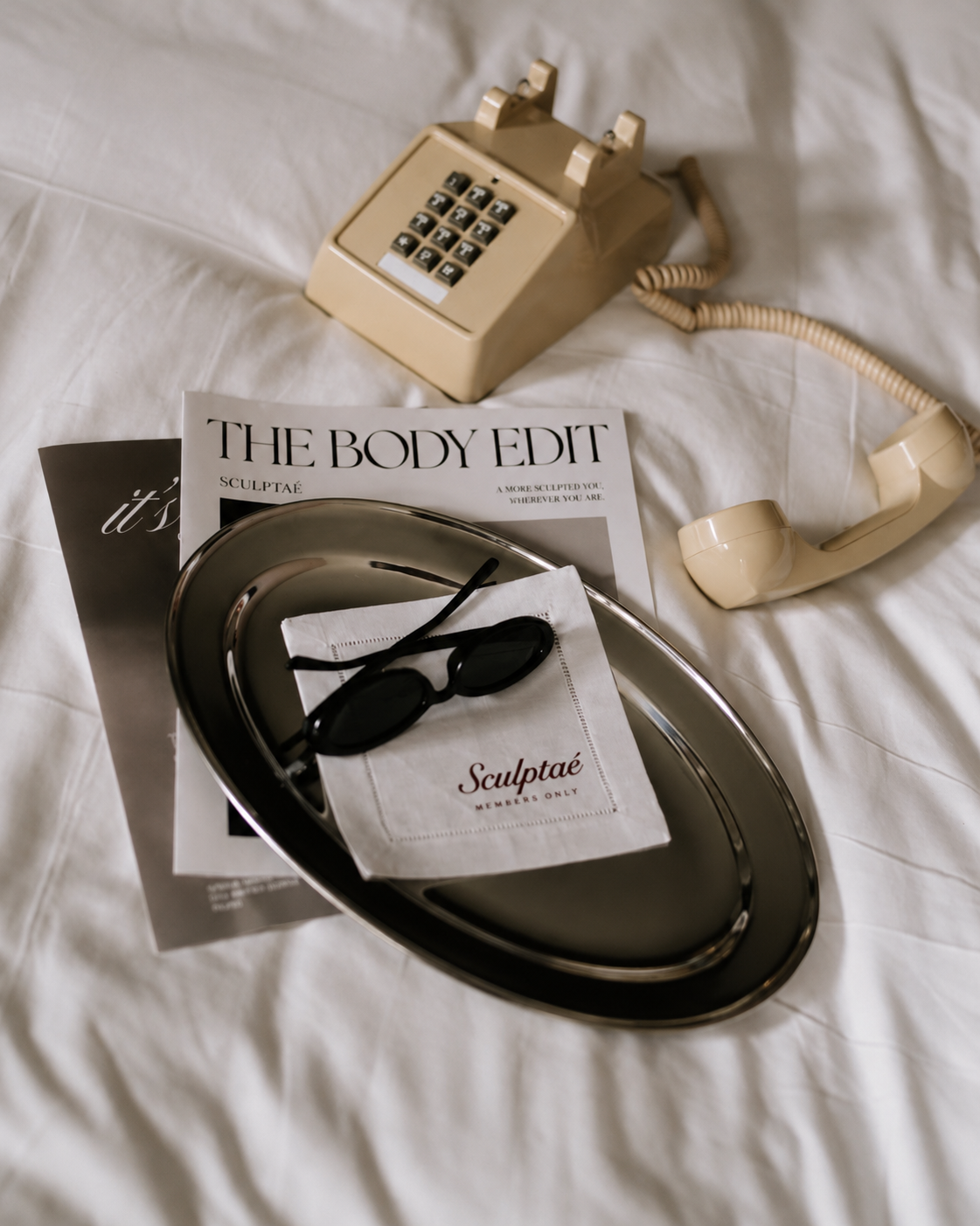 A vintage beige rotary telephone, a silver tray with black sunglasses and a white cloth with red text, a magazine titled 'The Body Edit,' and a pamphlet with partially visible text, all on a white bedsheet.