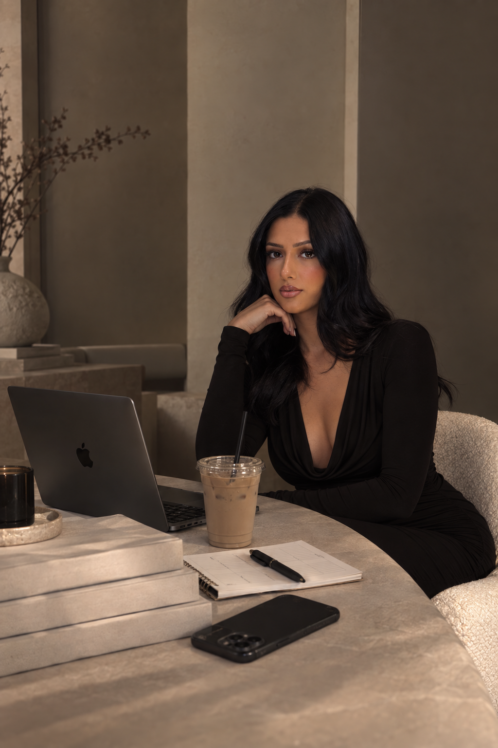 A woman with long black hair wearing a black dress sitting at a table with a laptop, iced coffee, smartphone, notebook, pen, and a black candle in a modern interior setting.