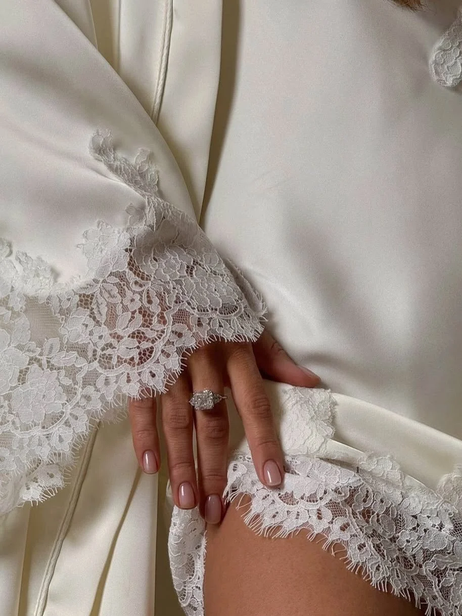 Close-up of a woman's hand with an engagement ring, resting on lace-edged satin fabric, possibly wedding dress.