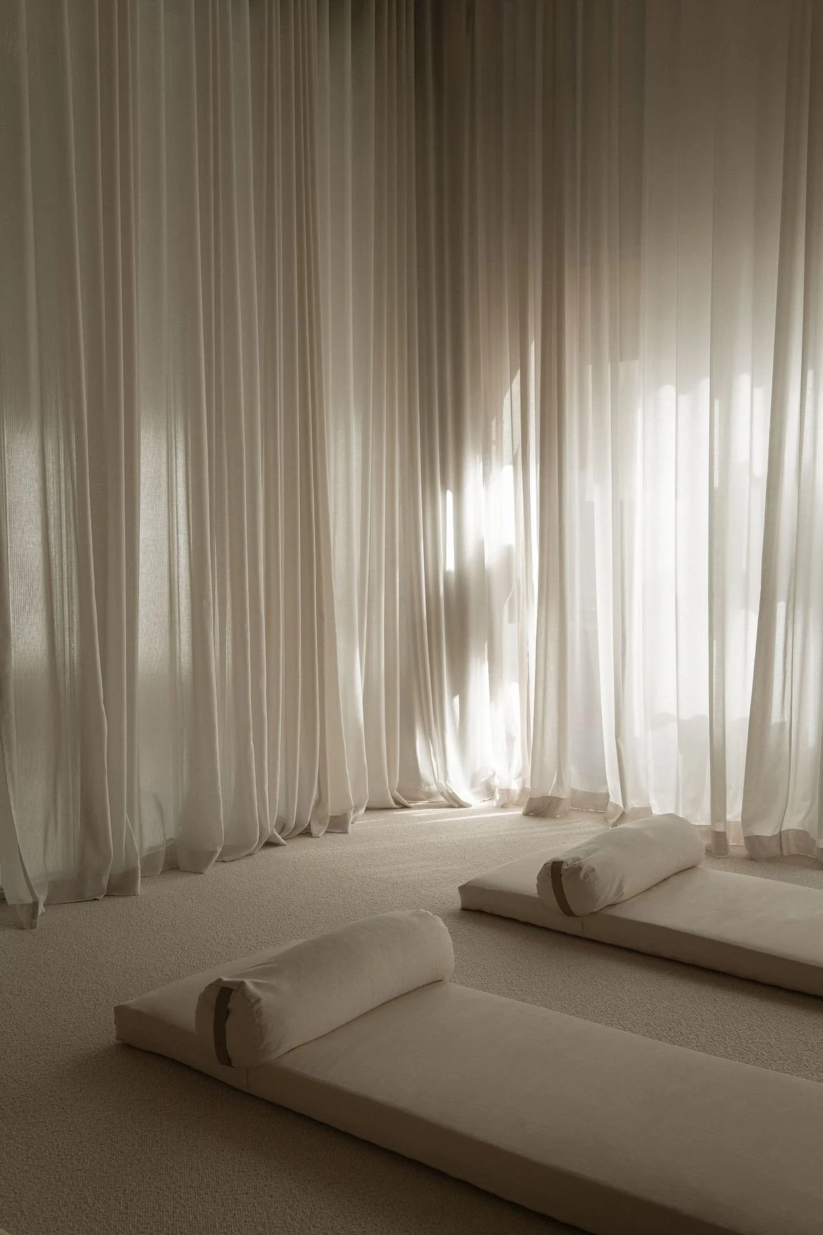 A room with white curtains covering large windows, and floor mats with pillows on a light-colored carpet.