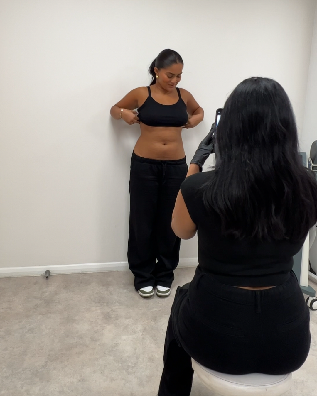 A woman is standing against a plain white wall, lifting her black sports bra to reveal her abdomen, while another woman takes a photo of her with a smartphone. The woman being photographed is dressed in black pants and white sneakers, with her hair tied back.
