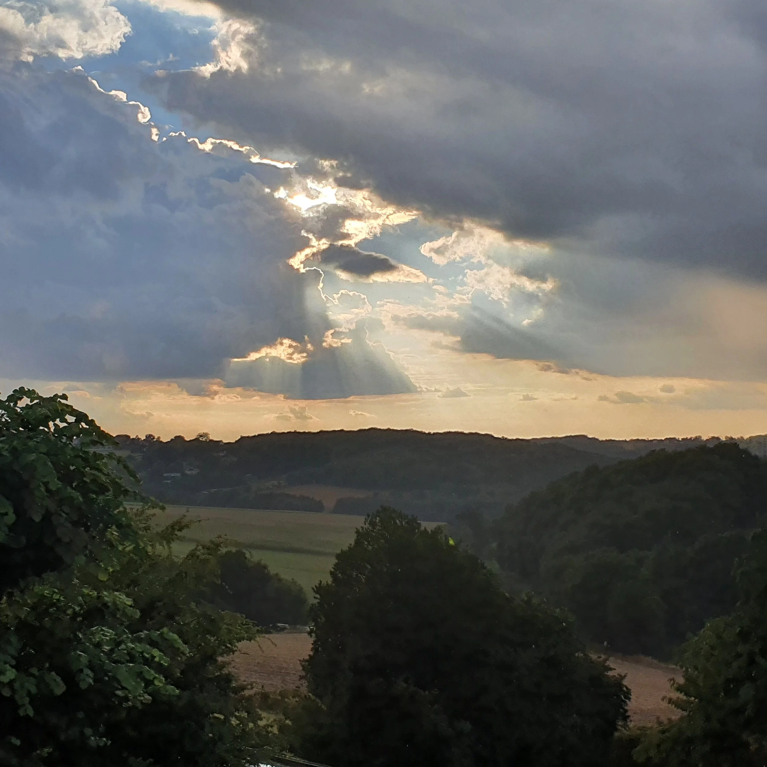 A scenic view of a cloudy sky with the sun peeking through the clouds and a silhouette of trees and hills in the distance.