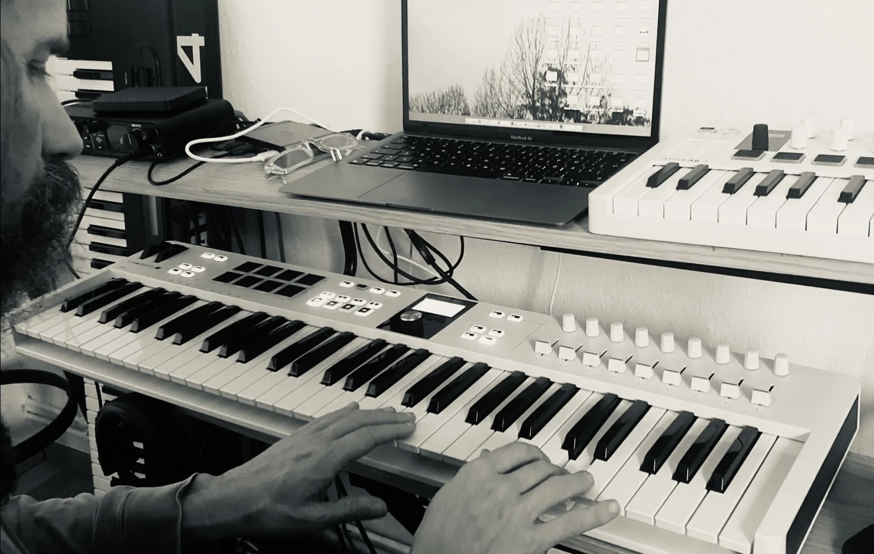 Close-up of a person playing a keyboard synthesizer, with a computer and MIDI controllers on a wooden desk in front of a wall.