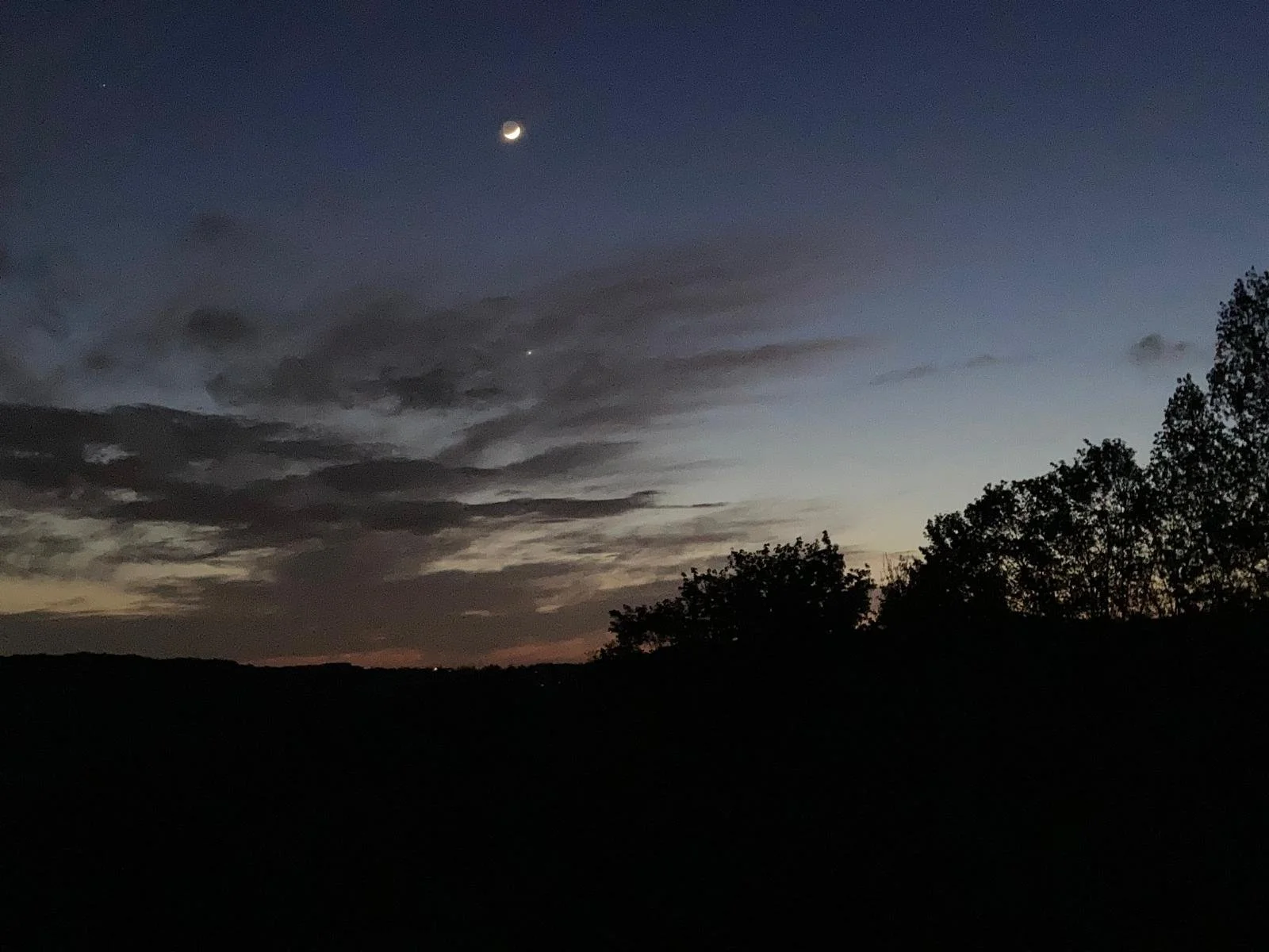 A landscape at dusk with a dark silhouette of trees and hills, a partly cloudy sky with dark clouds, a bright crescent moon, and a bright star or planet visible in the sky.