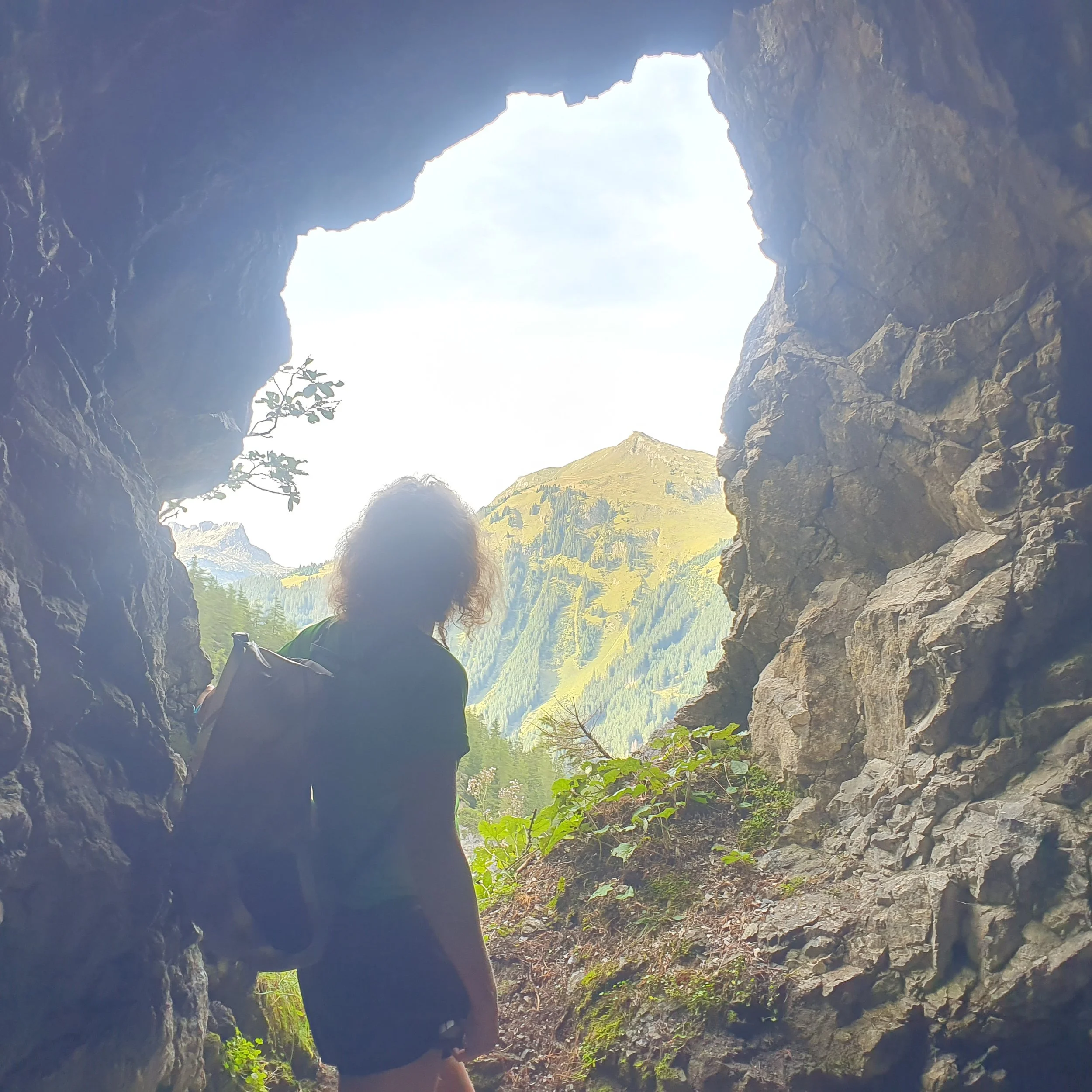 View from inside a cave opening looking out at green mountains and blue sky during daytime.