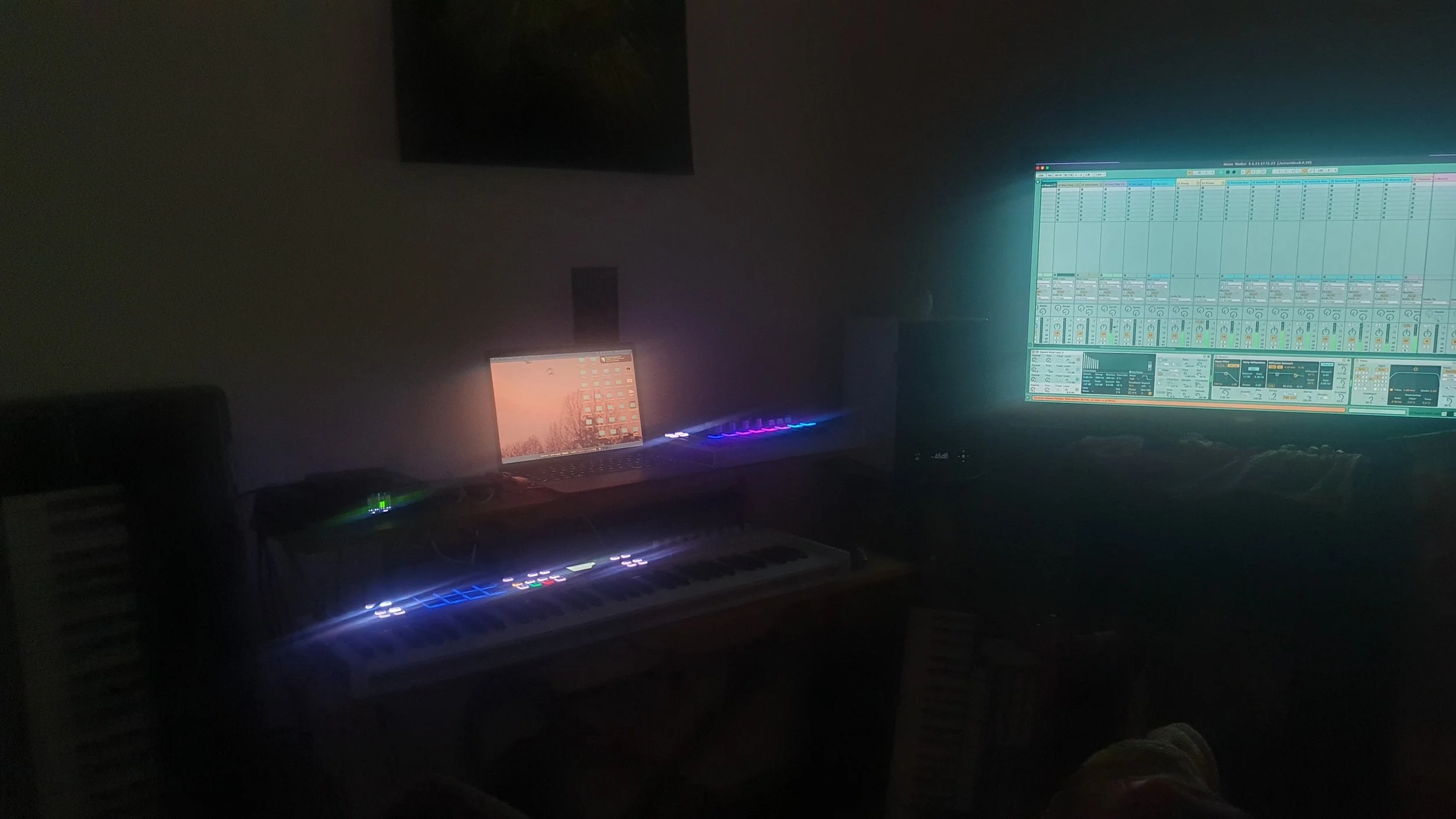 A music producer's setup in a dark room including a laptop, studio monitors, a MIDI keyboard, a large monitor displaying audio software, and some colorful LED lights.