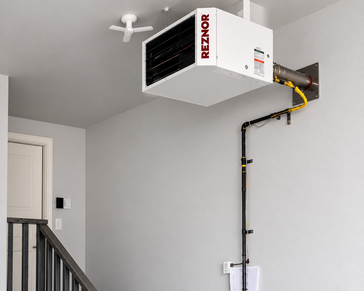 A room with a ceiling fan, a white HVAC unit labeled Reznor, and a black and yellow pipe running down the wall.