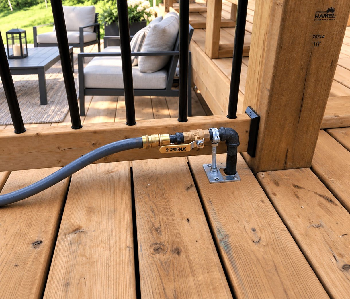 Close-up of a part of a wooden deck with a plumbing fitting attached to the wood, connected to a hose, and a valve handle. In the background, there are outdoor chairs and a table on the deck.