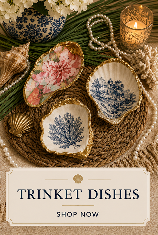 Decorative trinket dishes with floral and seaside motifs arranged on a woven mat with shells, pearls, a floral vase, and a lit candle for an elegant display.