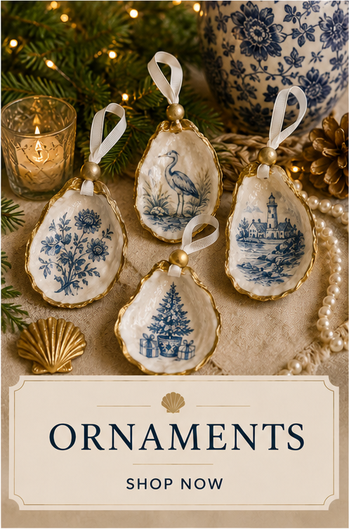 Decorative Christmas ornaments with gold edges and blue winter scenes, including a bird, lighthouse, decorated tree, and flowers, arranged on a beige surface with a candle, green pine branches, and a blue-and-white vase in the background.