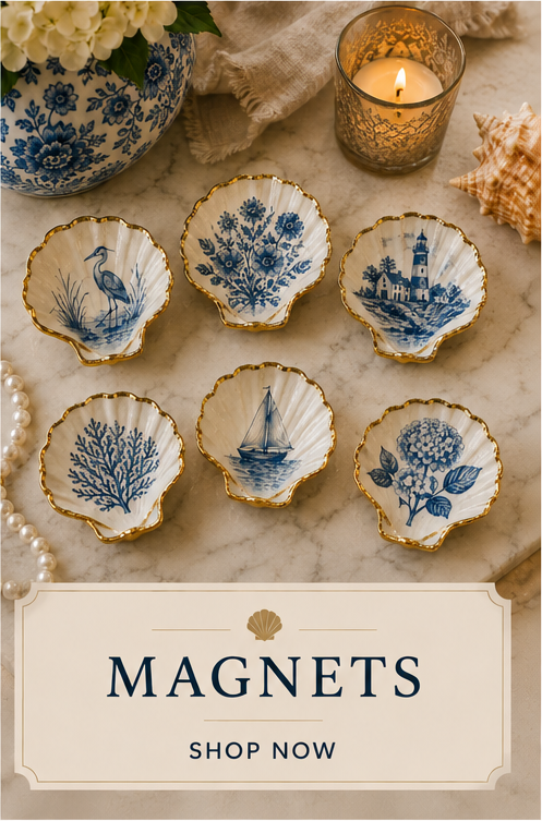 Six small decorative porcelain dishes with blue and white designs, featuring coastal and nautical themes, arranged on a marble surface with a candle, seashell, pearl necklace, and floral vase nearby.