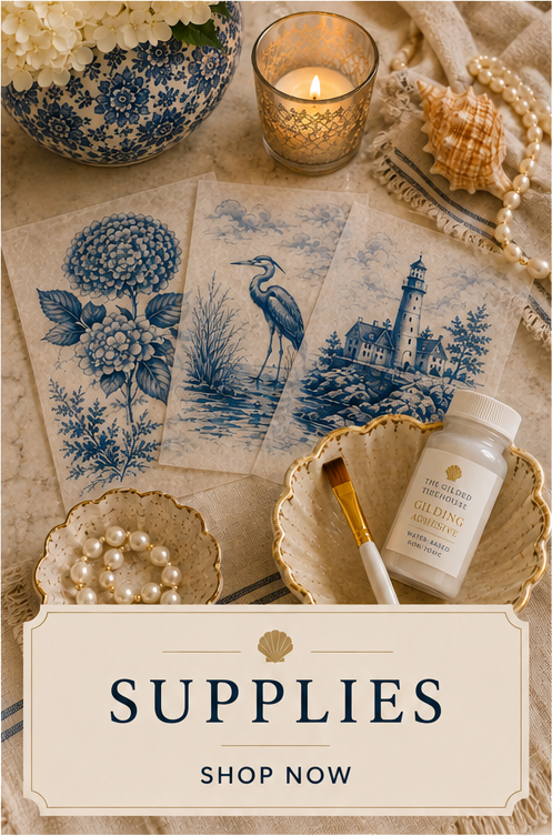 Decorative table setting with blue and white floral and landscape art prints, a lit candle in a patterned holder, seashells, pearls, a bottle of gilding adhesive, a paintbrush, and a sign saying 'Supplies Shop Now'.