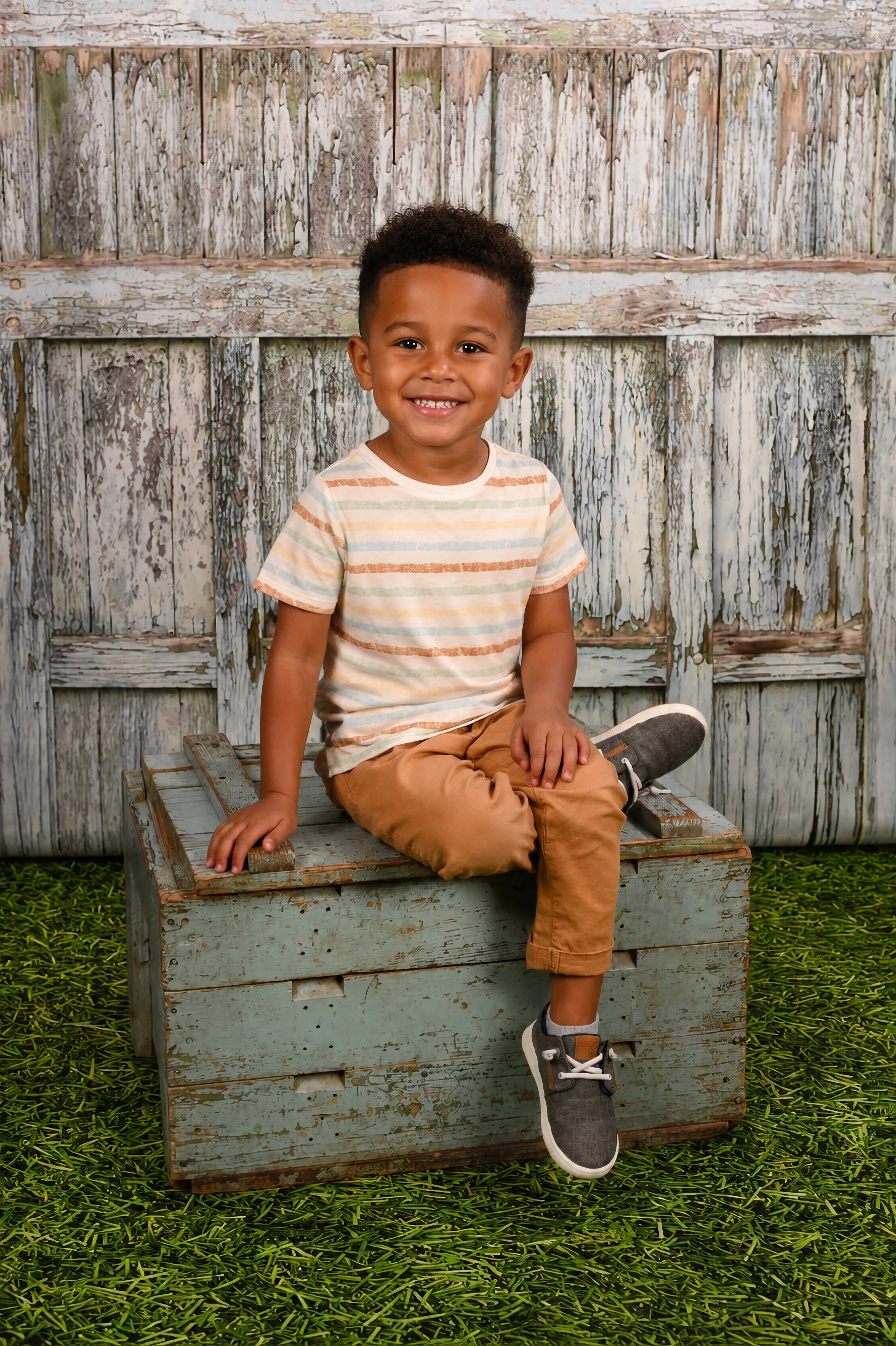 A young boy with curly hair and a big smile sitting on a rustic, weathered wooden bench against a backdrop of a distressed wood fence, with green grass beneath.