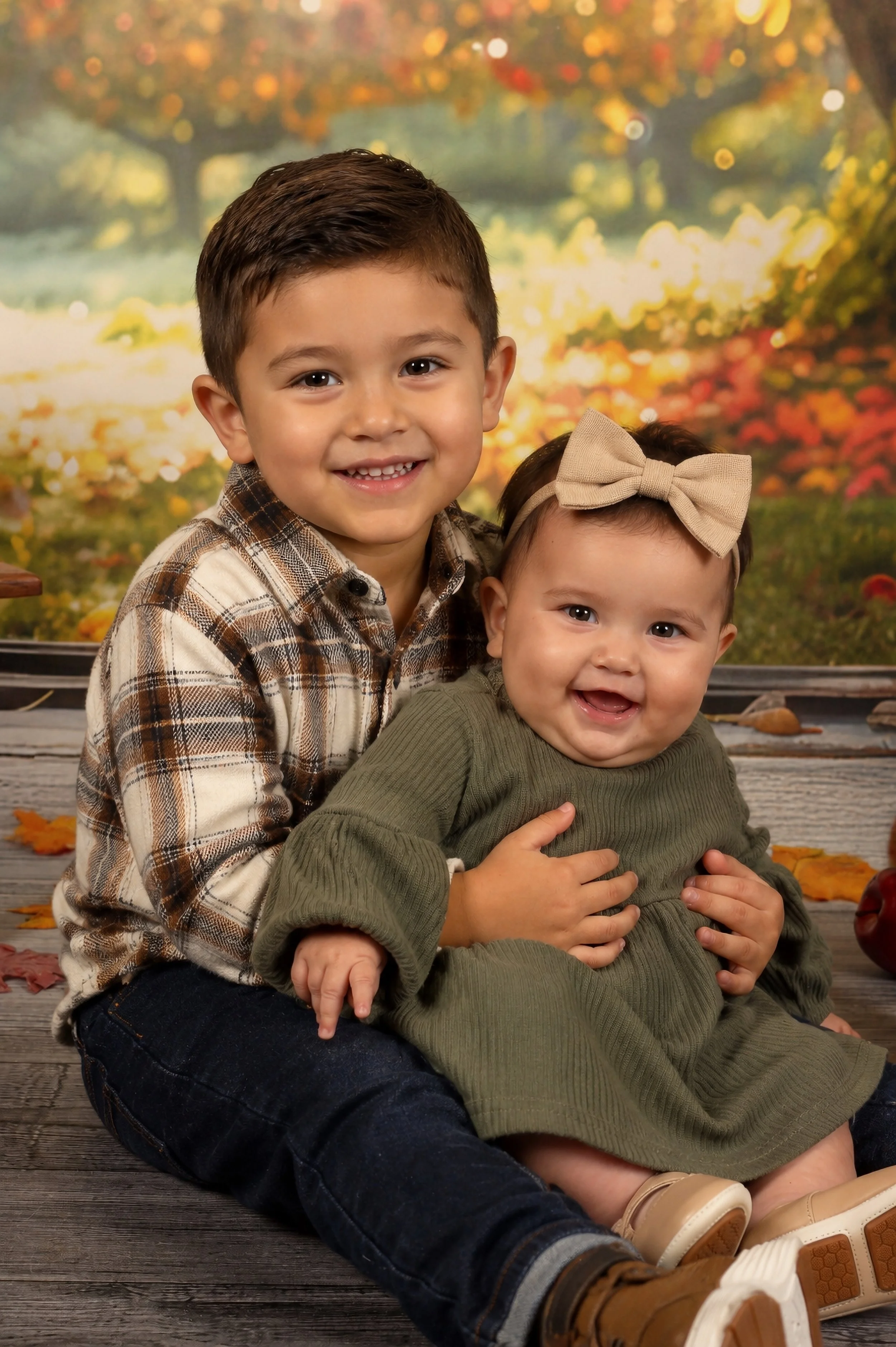 Two young children, a boy and a girl, sitting on a wooden floor with fall leaves. The boy has short dark hair, a schoolboy smile, and wears a plaid shirt. The girl has dark hair with a bow, a big smile, and wears a green sweater.
