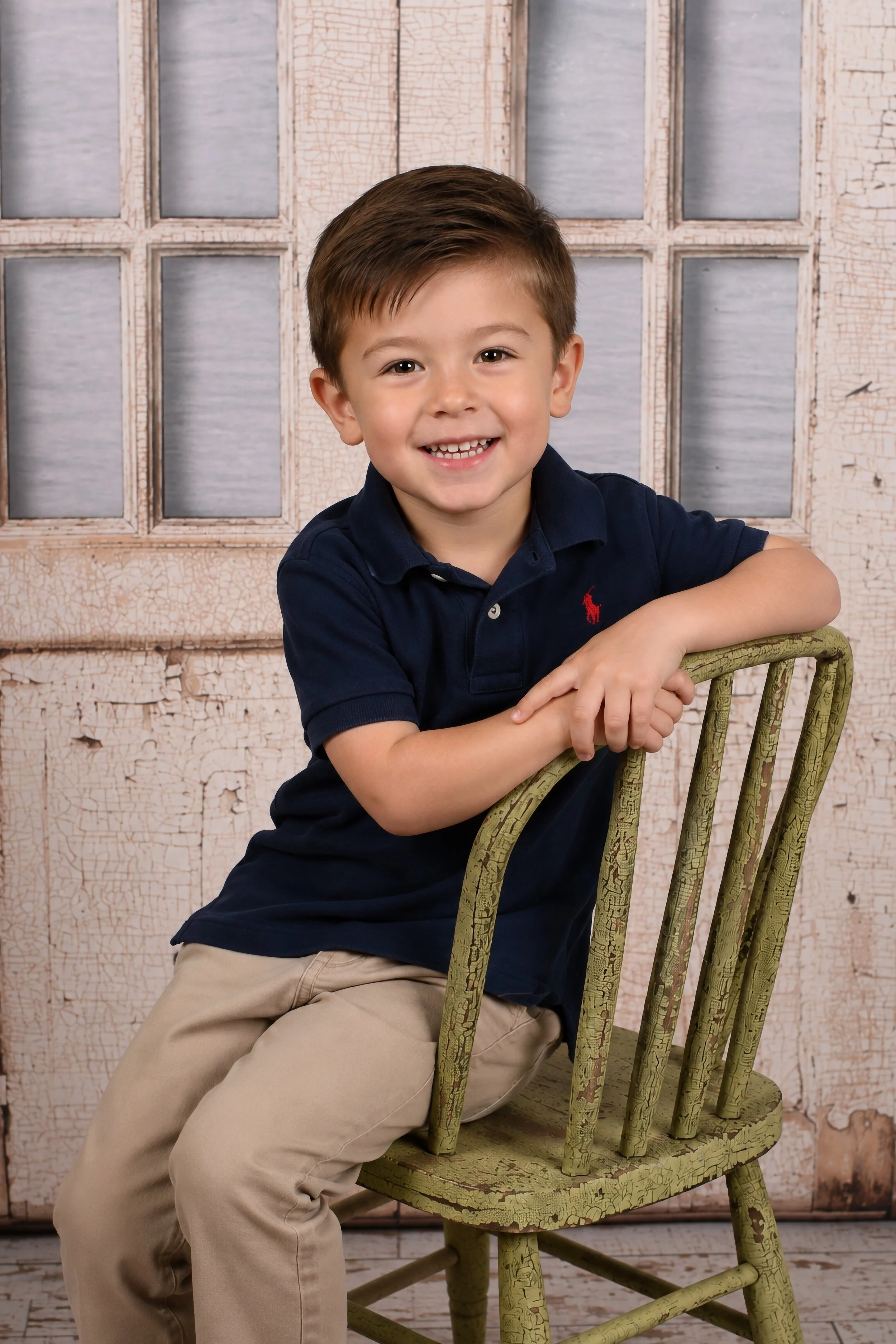 A young boy with brown hair, smiling and wearing a navy polo shirt and khaki pants, sitting on a green wooden chair in front of a rustic, peeling white-paneled wall.