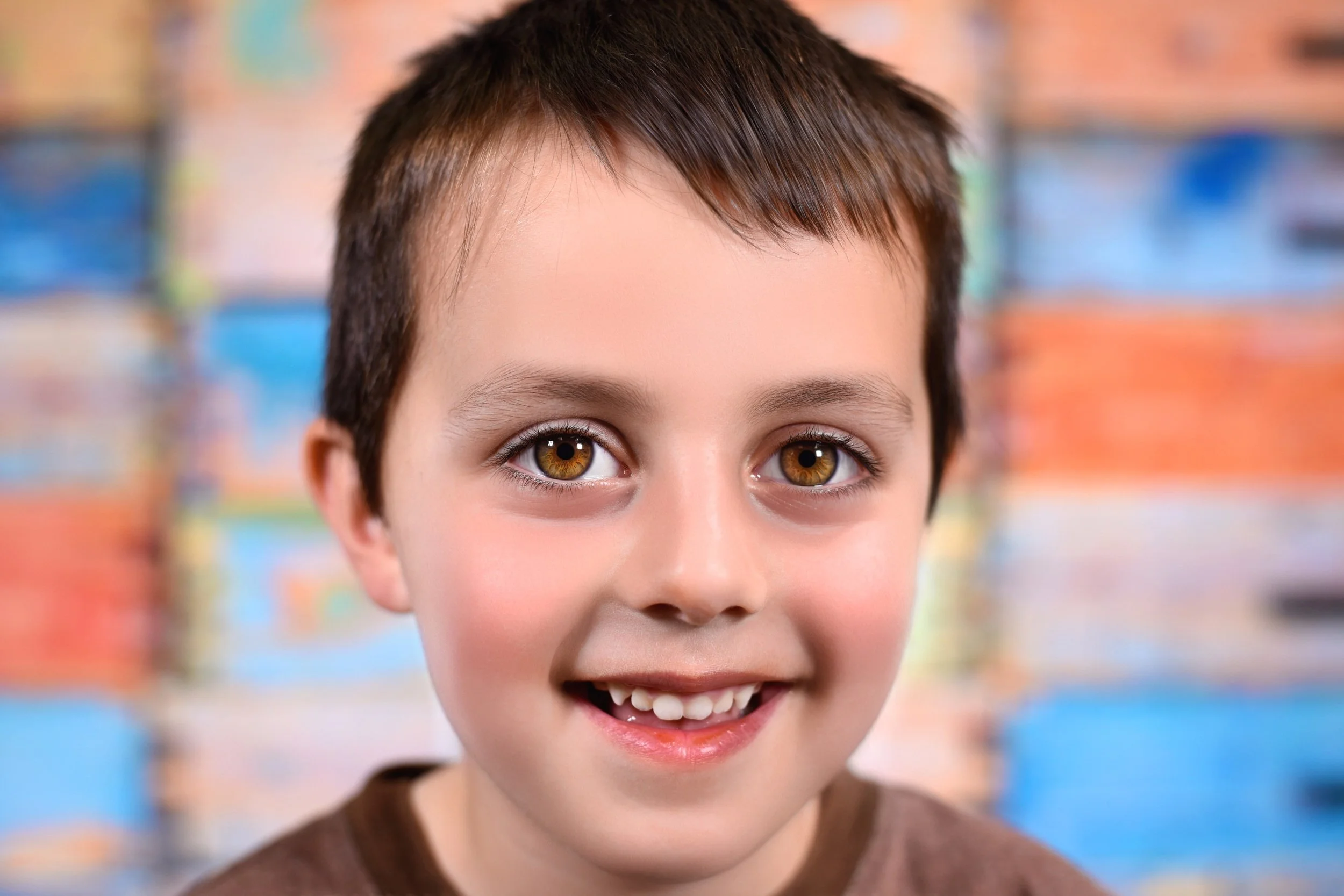 Close-up of a young boy smiling, showing his eyes and part of his teeth, with a blurred colorful background.