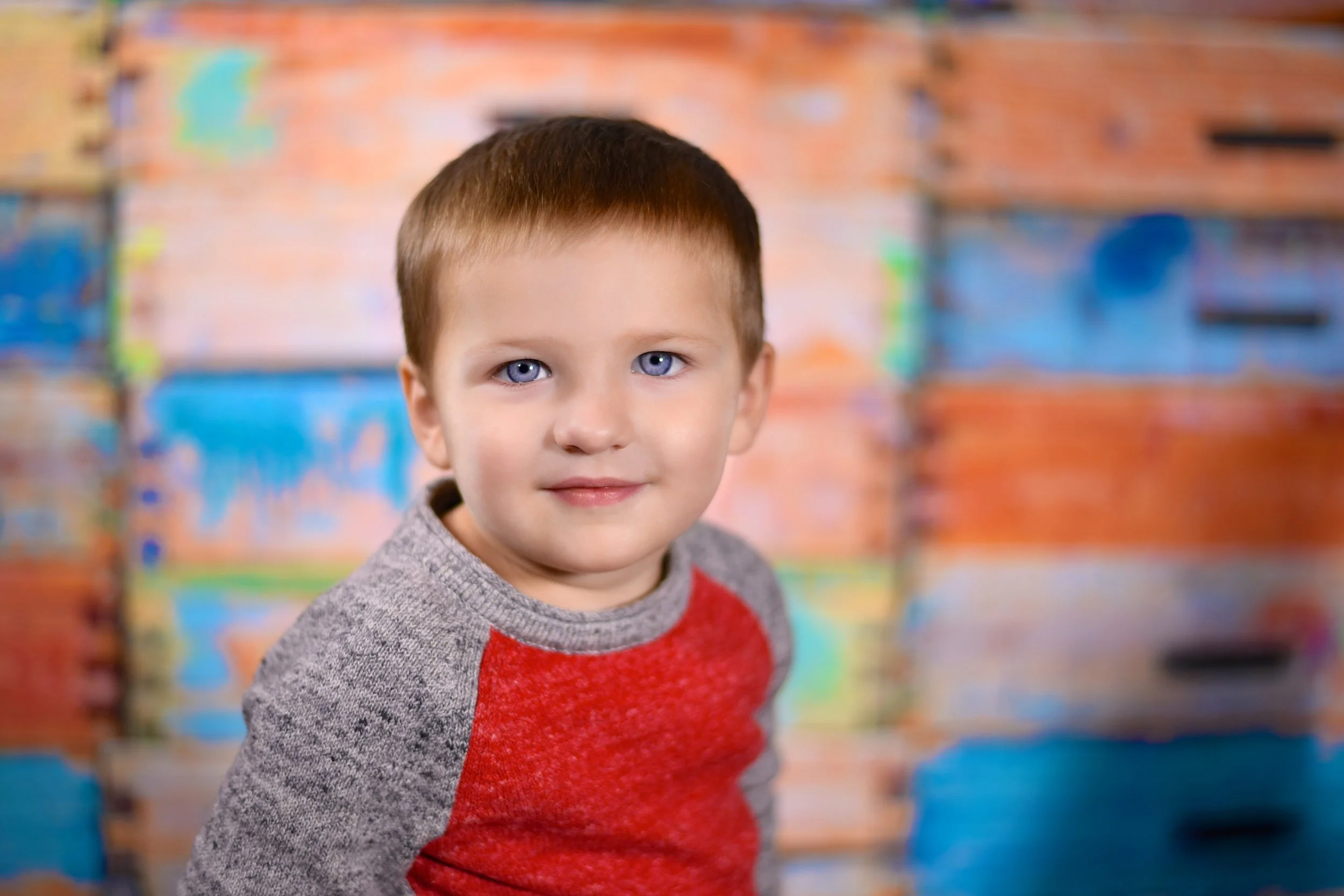 Close-up of a young boy with short brown hair and blue eyes wearing a gray and red sweater, standing in front of a colorful, abstract background.